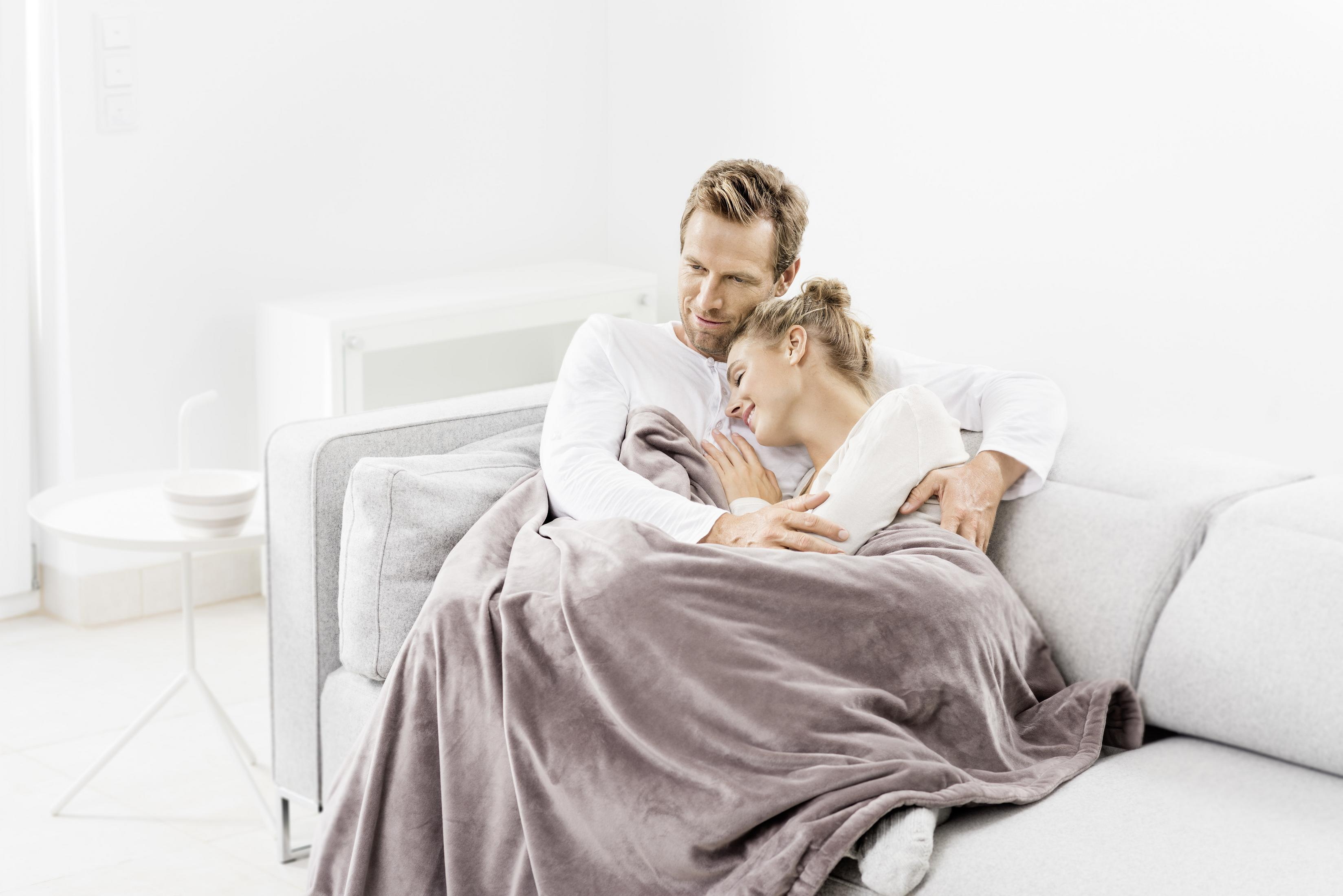 A couple cuddles under a blanket on a gray sofa. The background is a white wall and a cabinet.