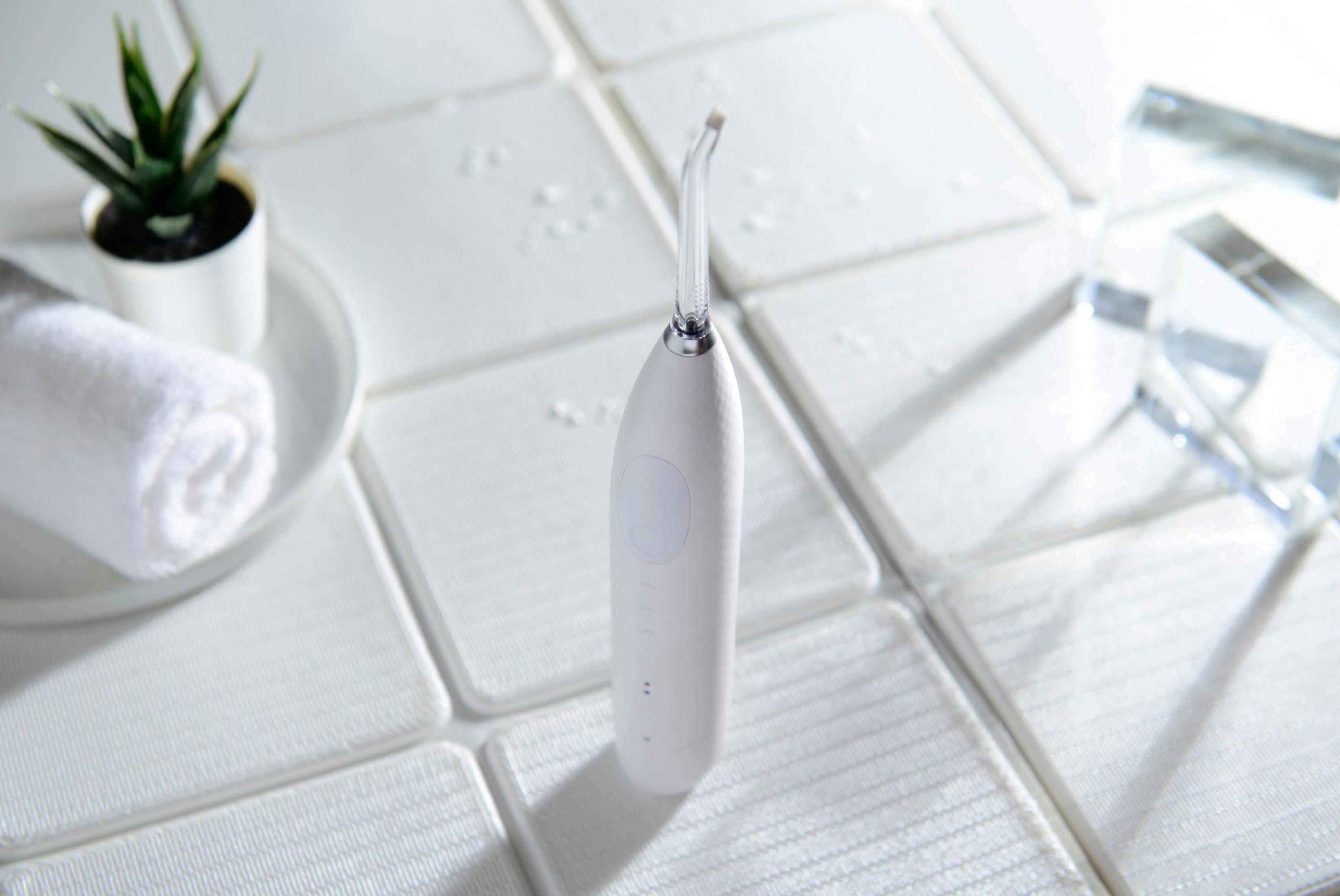 Biały irygator do jamy ustnej na białej kafelkowej powierzchni z małą rośliną i ręcznikiem w tle.