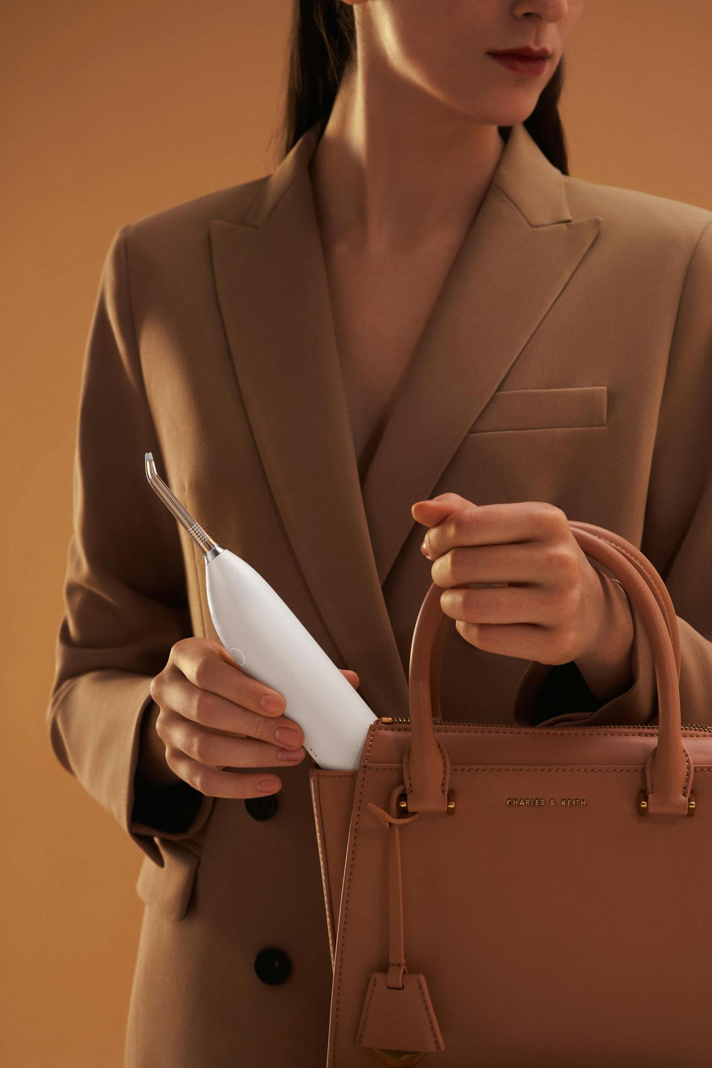 Kobieta w brązowej marynarce trzyma w torebce biały irygator do jamy ustnej, na brązowym tle.
