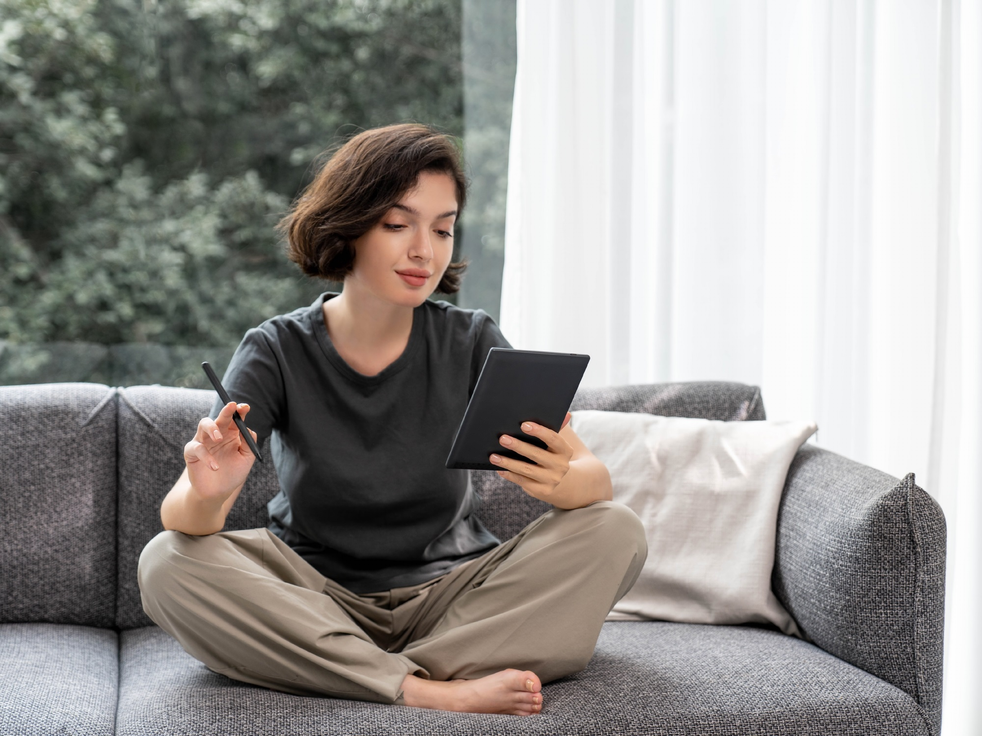 Eine Frau sitzt auf einem grauen Sofa und hält ein Tablet und einen Stift. Sie ist in einem hellen Raum.