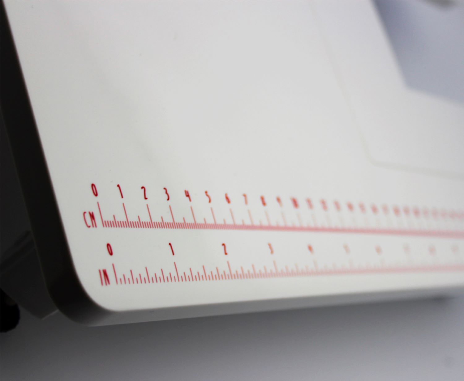 Dispositivo de medición blanco con marcas rojas de cm/pulgadas sobre fondo blanco.
