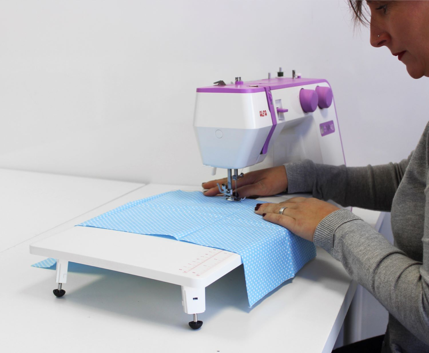 Una mujer cose tela azul en una máquina de coser blanca sobre una mesa blanca. El fondo es blanco.