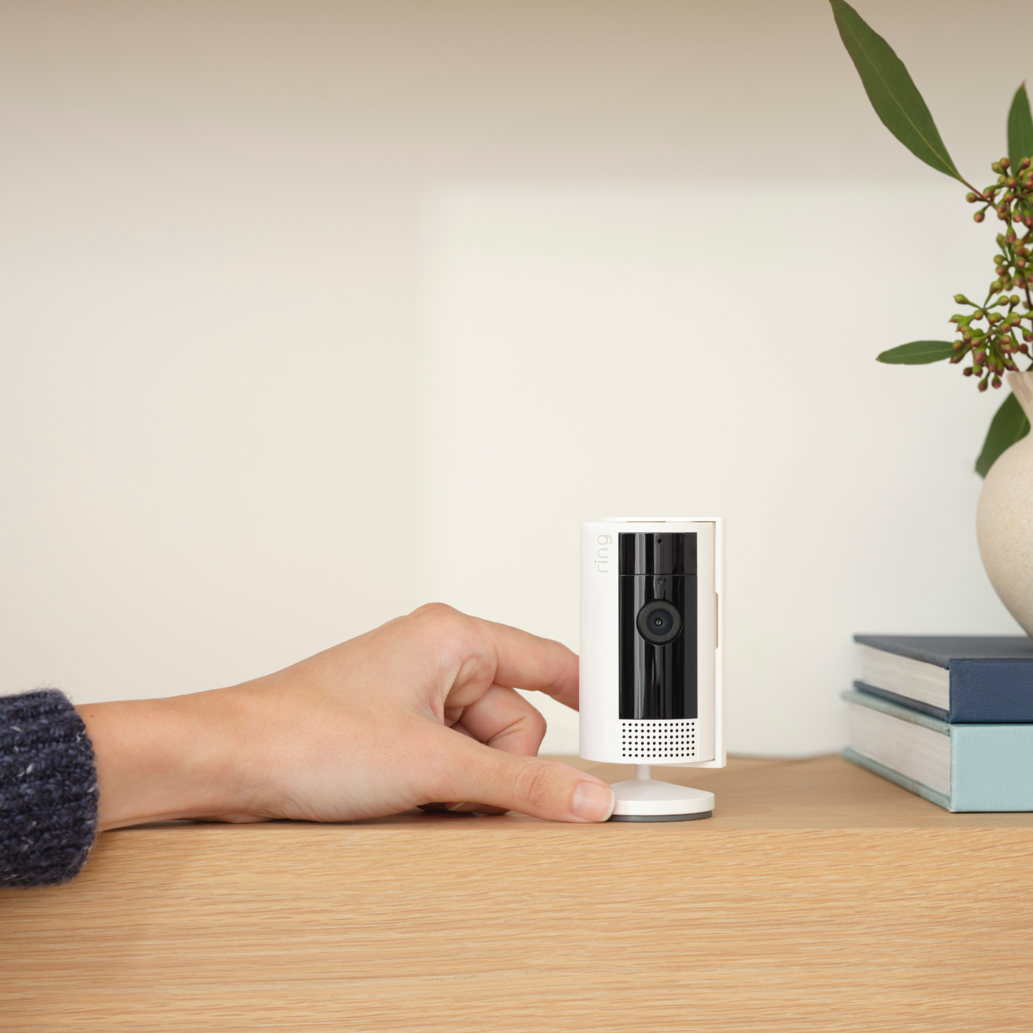 Ręka kobiety sięgająca po białą kamerę bezpieczeństwa Ring na drewnianej powierzchni. Książki w pobliżu.