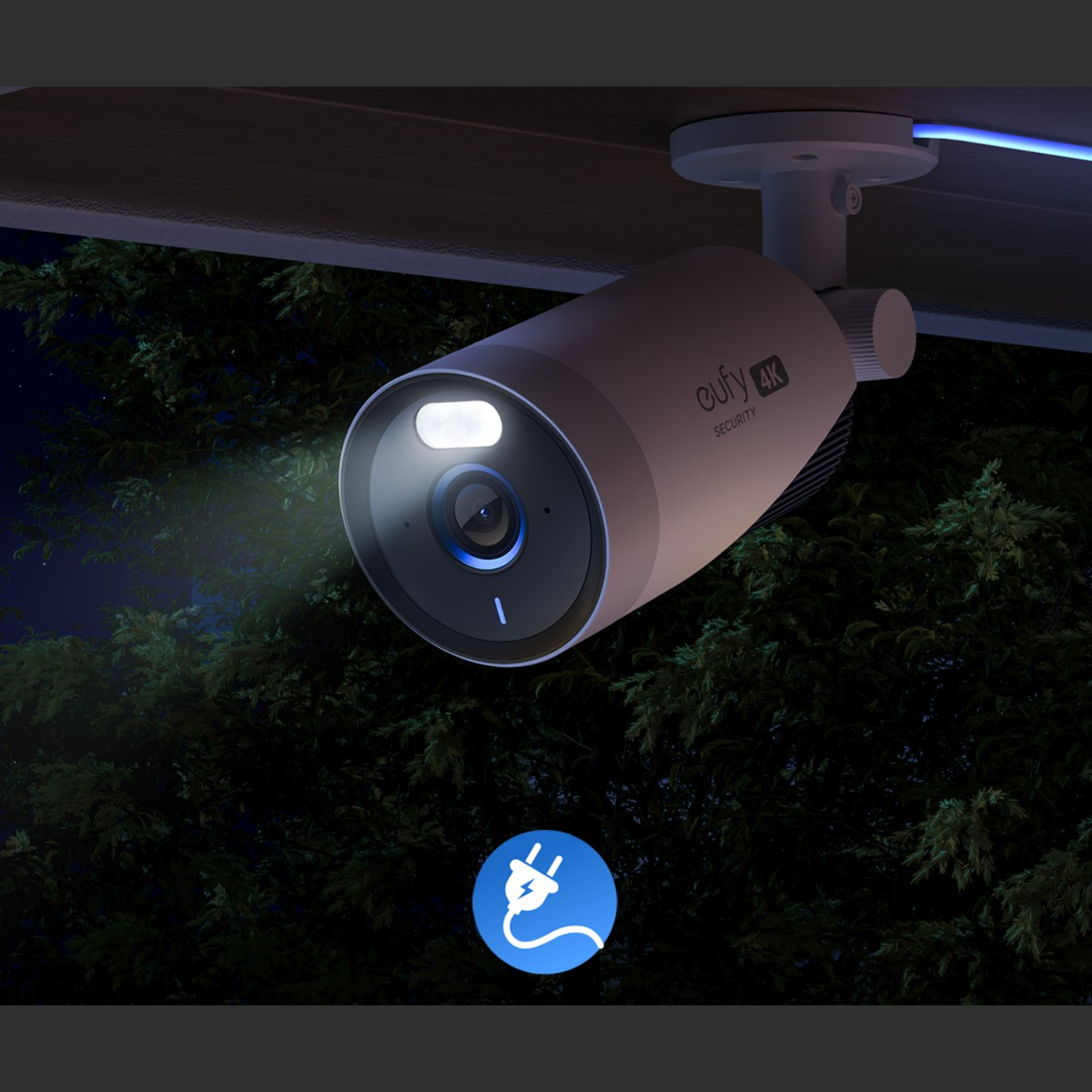 Zewnętrzna kamera bezpieczeństwa z włączonym światłem, zamontowana pod dachem. W tle widoczne drzewa.