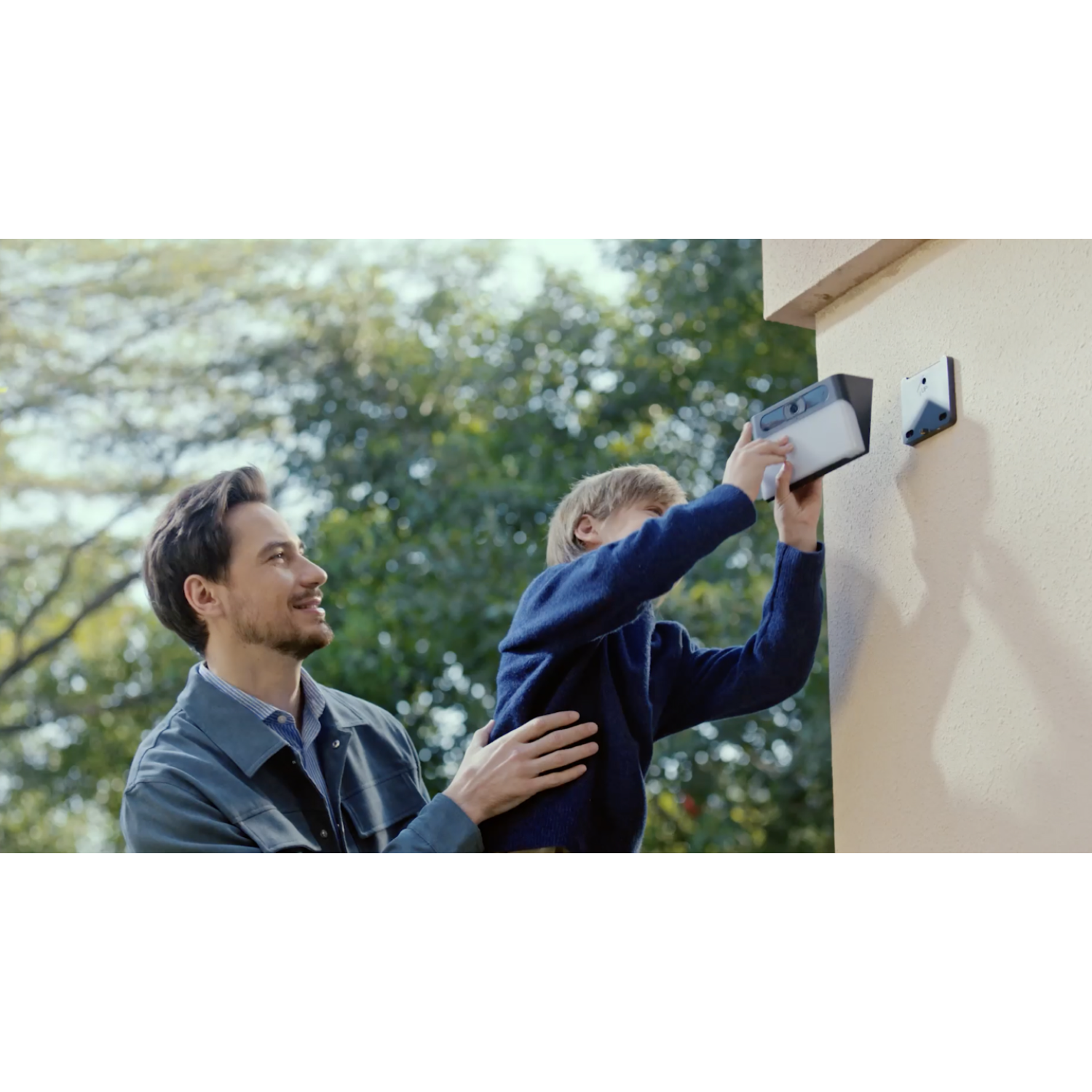 Mężczyzna i chłopiec instalują kamerę bezpieczeństwa na beżowej ścianie. Mężczyzna trzyma chłopca.