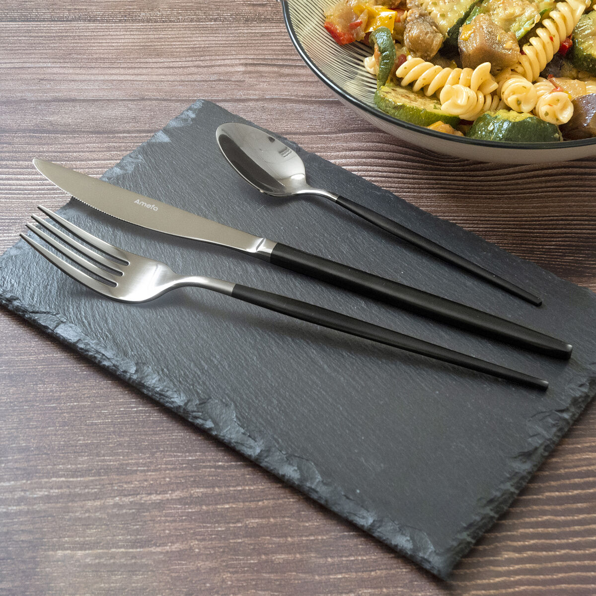 Forchetta, coltello e cucchiaio su una lavagna nera, con una ciotola di cibo sullo sfondo su un tavolo di legno.