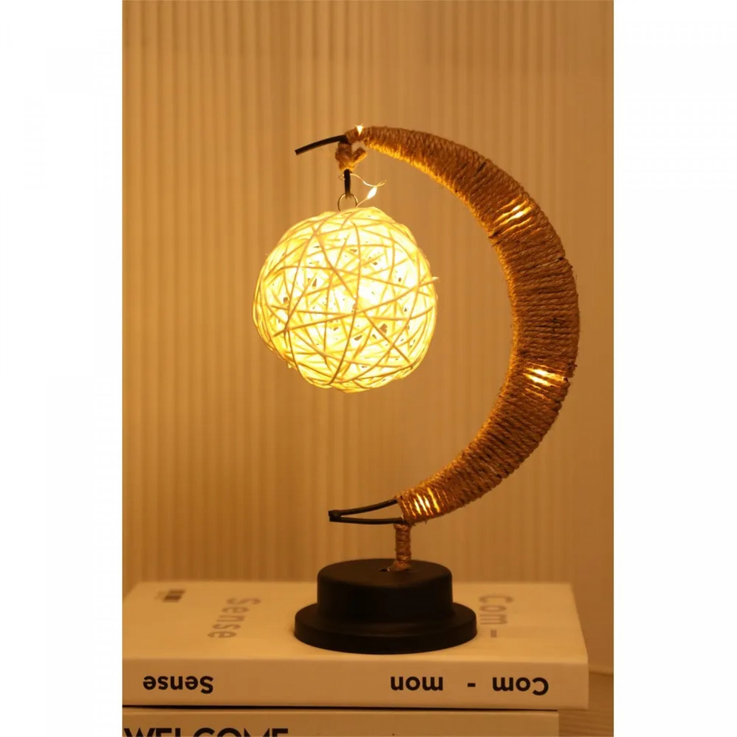 Mondförmige Lampe mit gewebter Kugel, auf einem Bücherstapel. Warmes Licht beleuchtet die Szene.