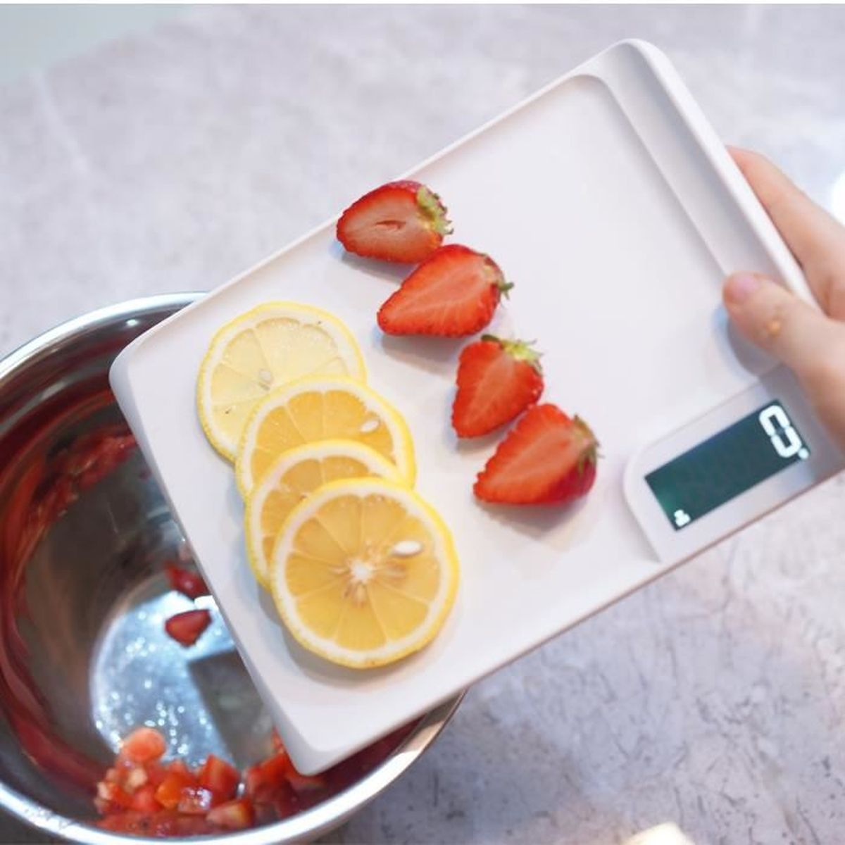 Eine weiße Waage mit aufgeschnittenen Zitronen und halbierten Erdbeeren, mit einer Schüssel mit gehackten Tomaten im Hintergrund.