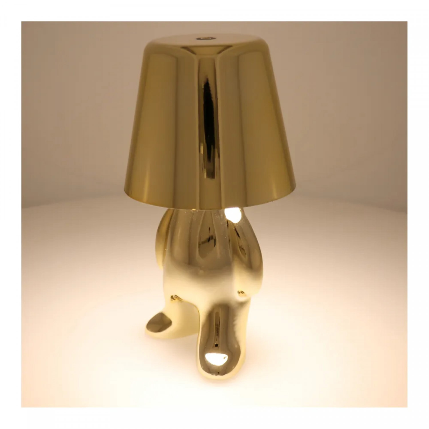 Goldene Lampe in Form einer Person, nach vorne gebeugt. Die Lampe steht auf weißem Grund und strahlt Licht aus.