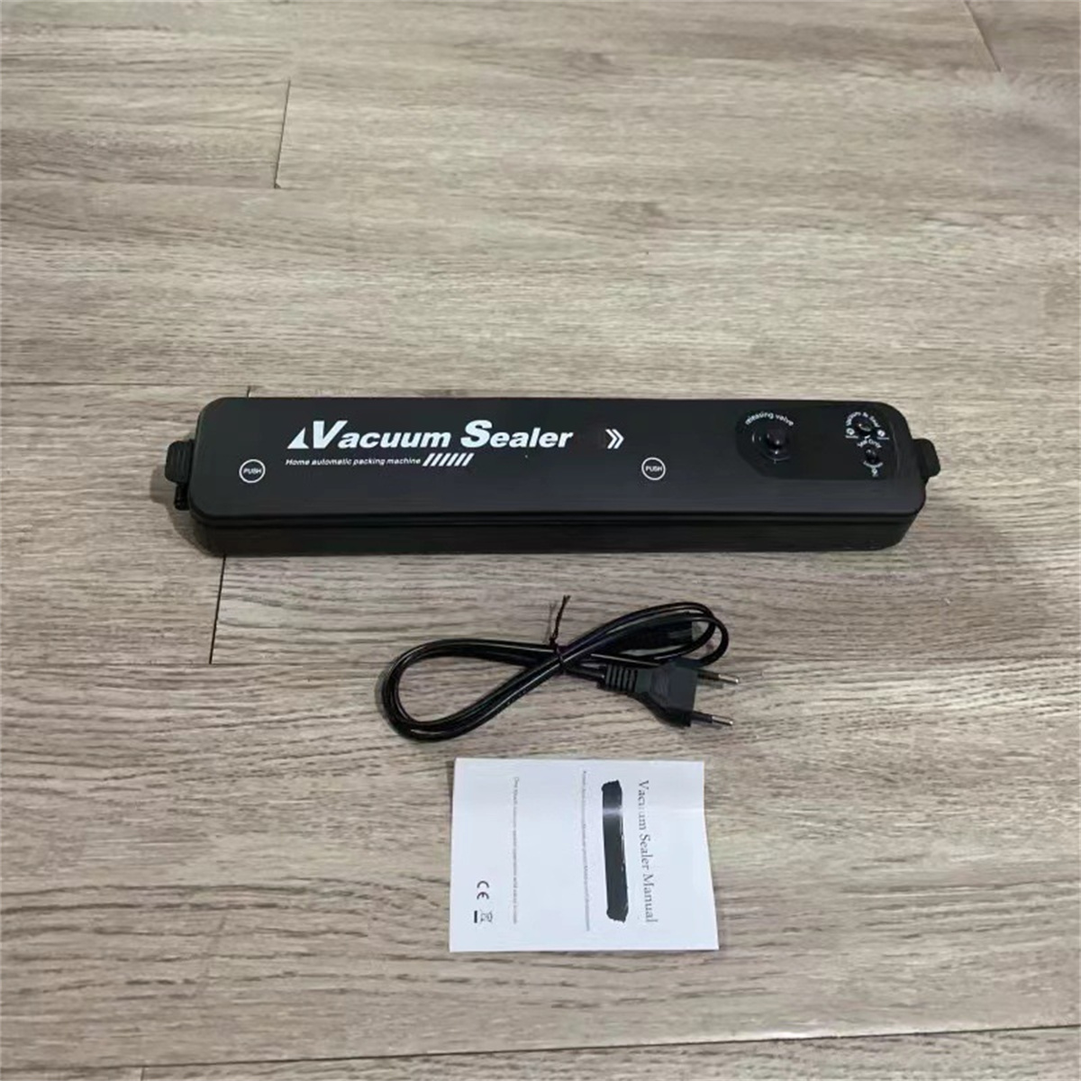 Ein schwarzes Vakuumiergerät, ein Netzkabel und eine Bedienungsanleitung liegen auf einer Holzoberfläche.