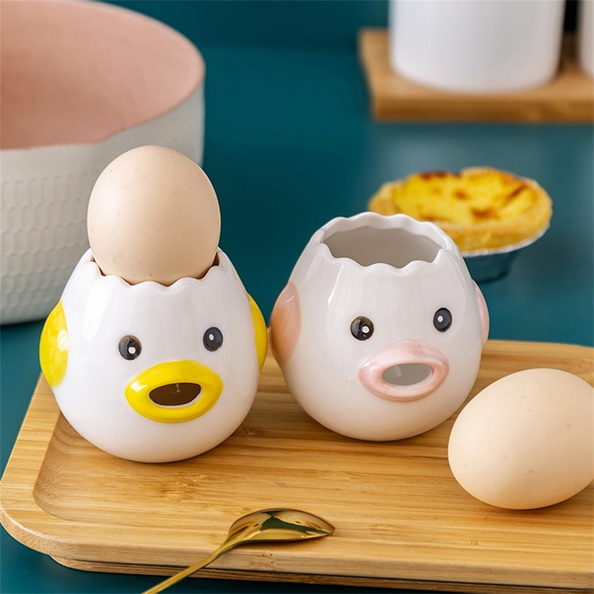 Zwei keramische Eiertrenner in Kükenform und Eier auf einem Holztablett.