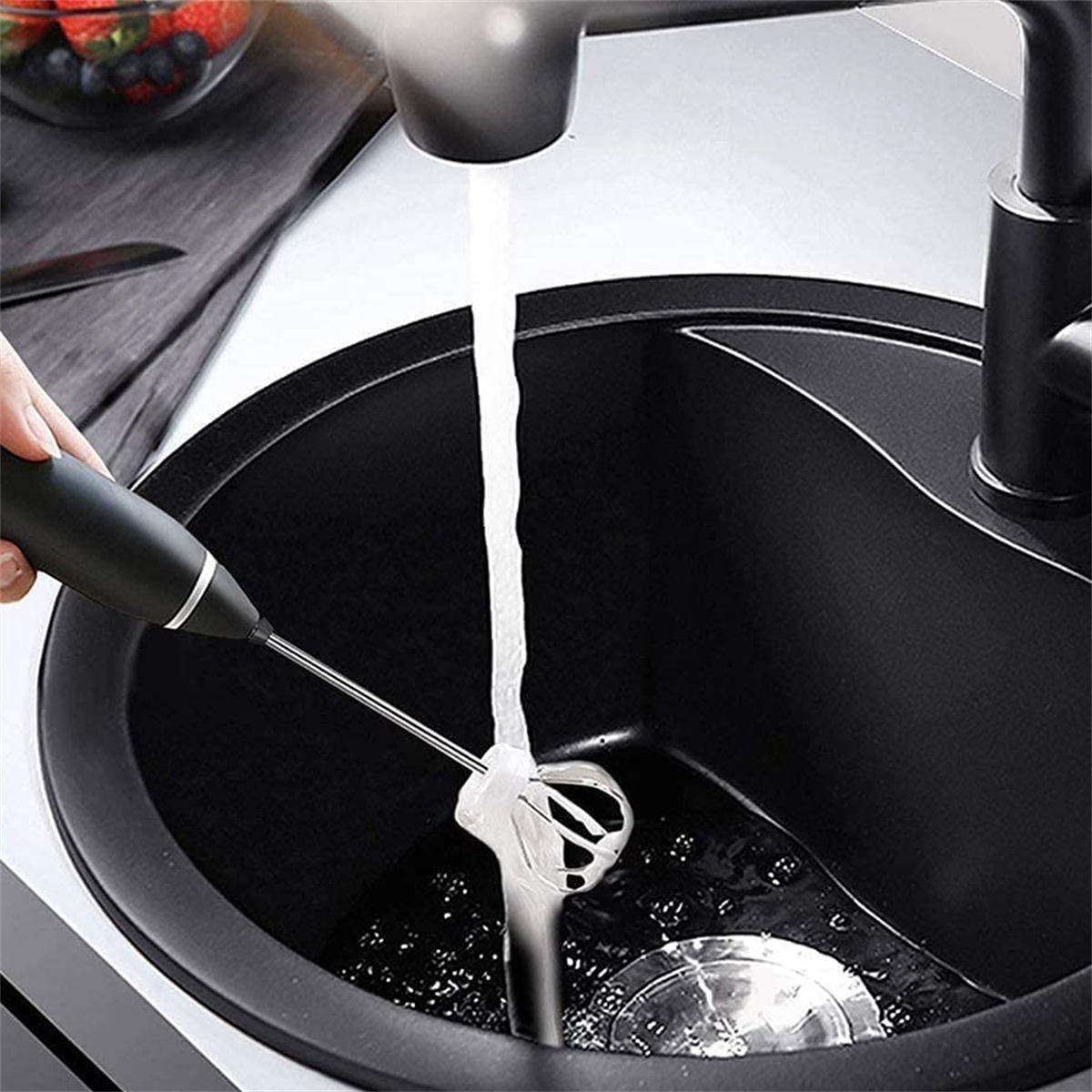 Hand mit Milchaufschäumer in der Spüle, Wasser läuft, schwarzes Spülbecken sichtbar.