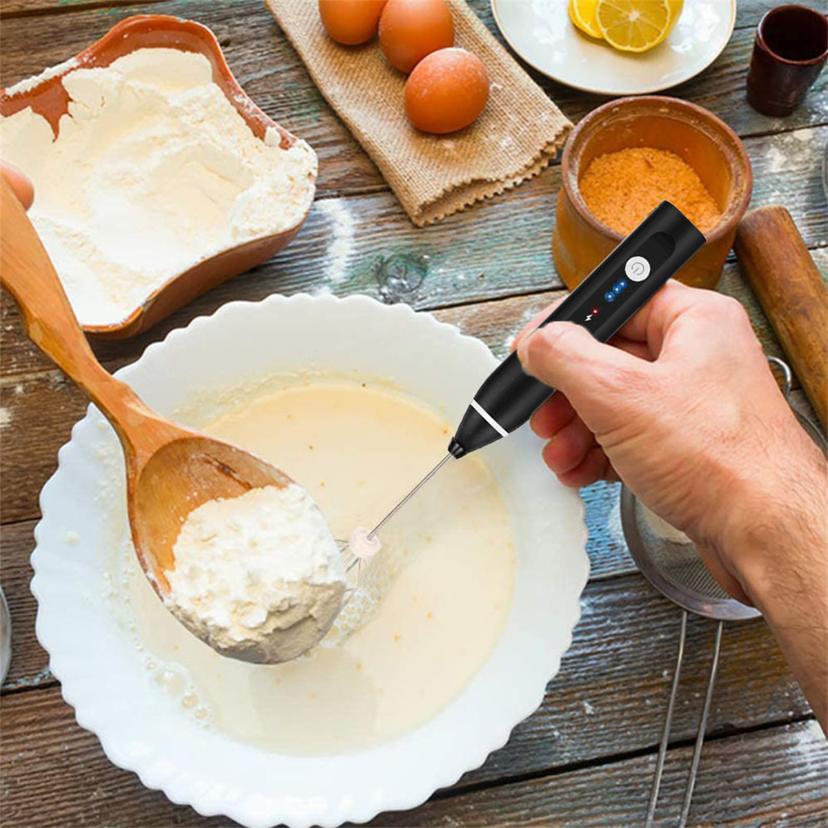 Eine Hand benutzt einen Stabmixer, um Teig in einer weißen Schüssel zu verrühren. Zutaten und Eier sind im Hintergrund.