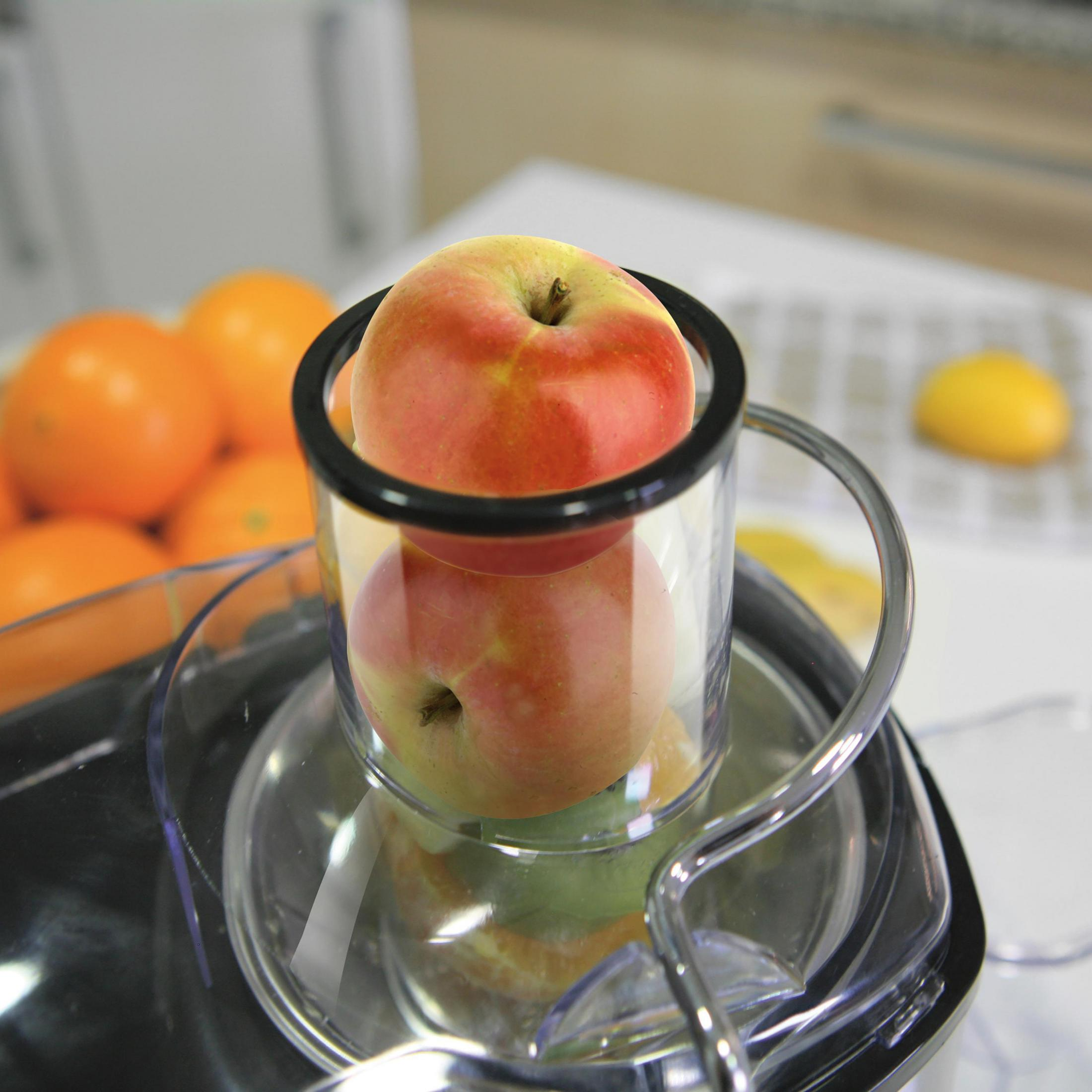 Dos manzanas dentro de un exprimidor transparente, con naranjas y un limón al fondo.