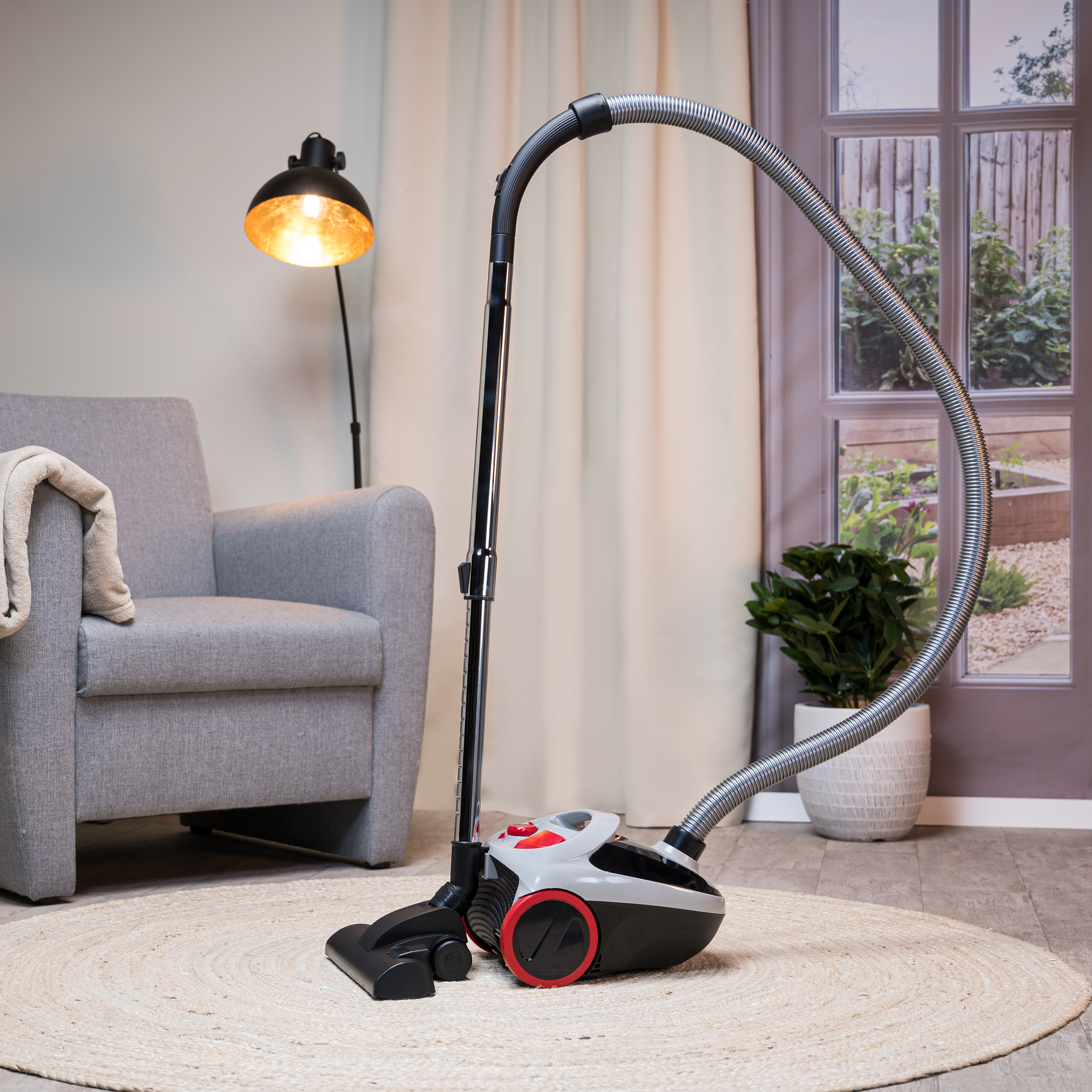 Un aspirateur est posé sur un tapis. Il est noir, gris et rouge. Une lampe et une chaise en arrière-plan.