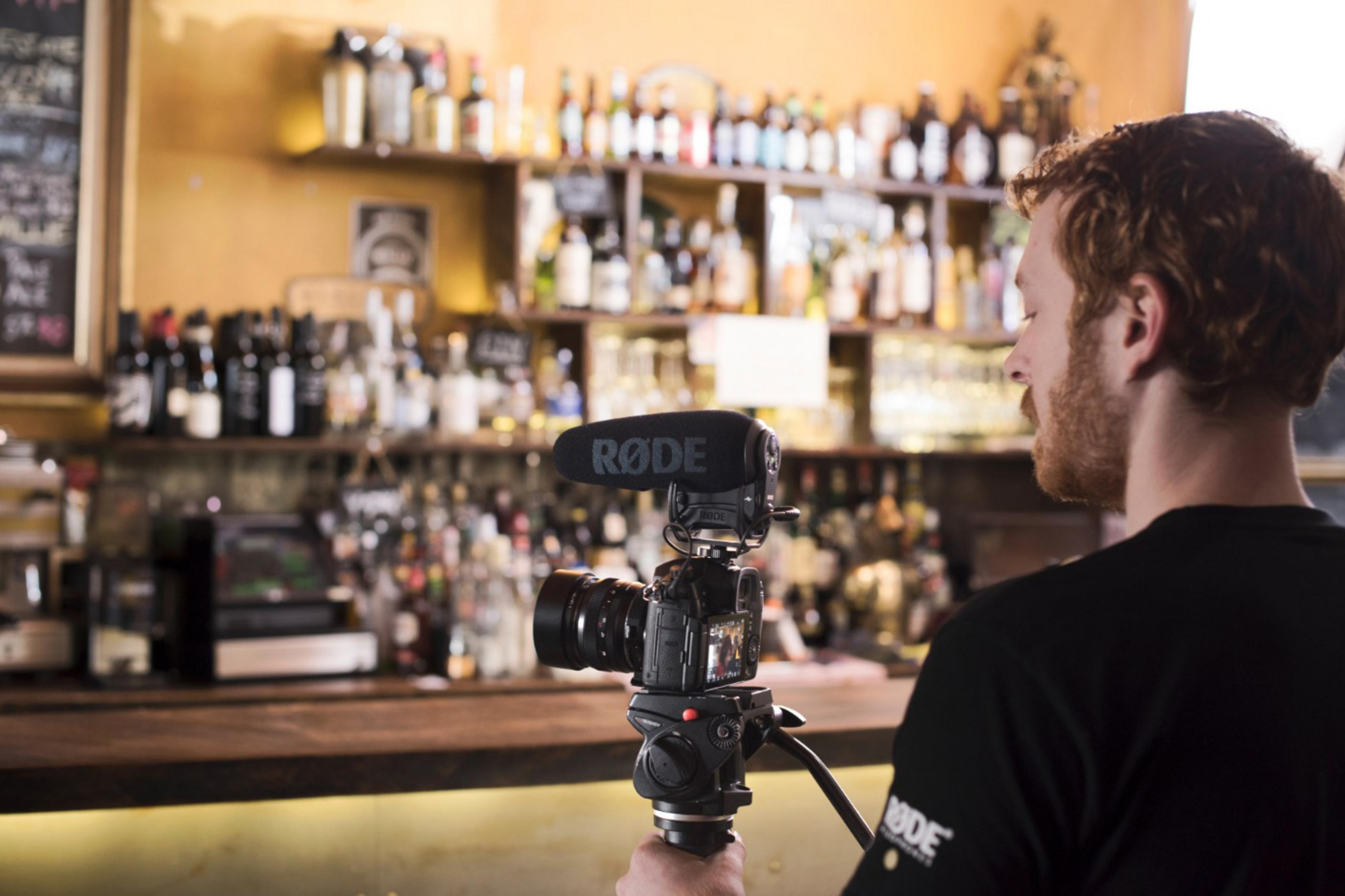 Mężczyzna filmujący kamerą i mikrofonem w barze, z butelkami na półce.