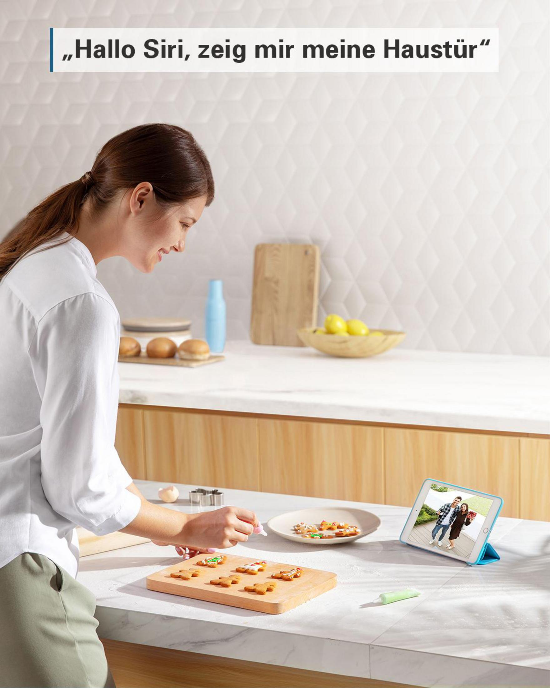 Kobieta w kuchni z tabletem pokazującym wideo. Dekoruje ciasteczka. Tekst: Cześć Siri, pokaż mi moje drzwi wejściowe.