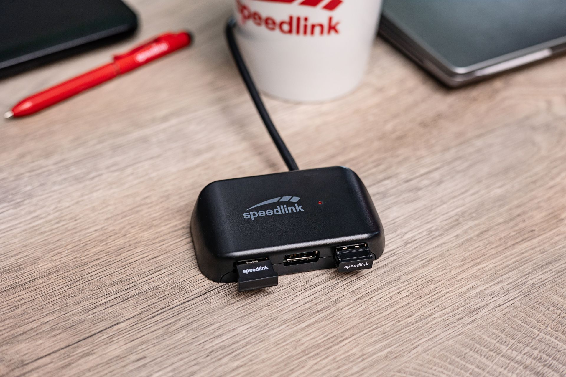 Un hub USB nero con due porte, una luce rossa e un logo Speedlink, posizionato su un tavolo di legno.
