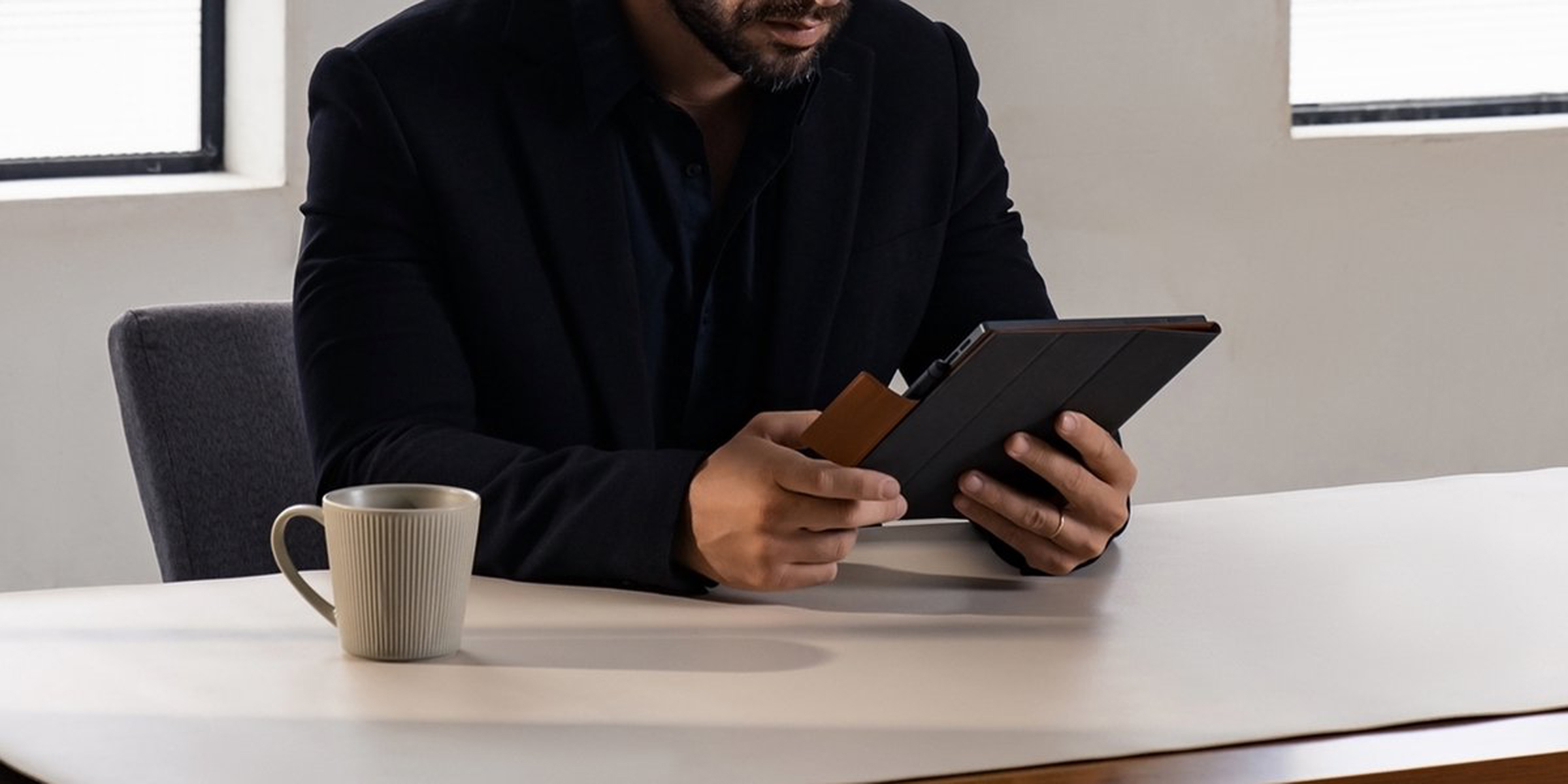 Person sitzt an einem Tisch und betrachtet ein Tablet. Auf dem Tisch befinden sich eine Tasse und eine Karte.