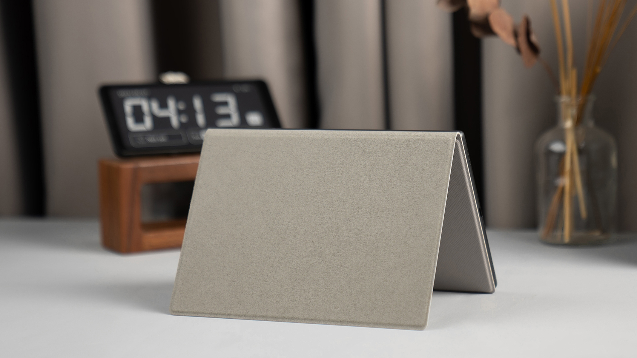 Custodia grigia per tablet appoggiata su un tavolo con un orologio e una pianta sullo sfondo.
