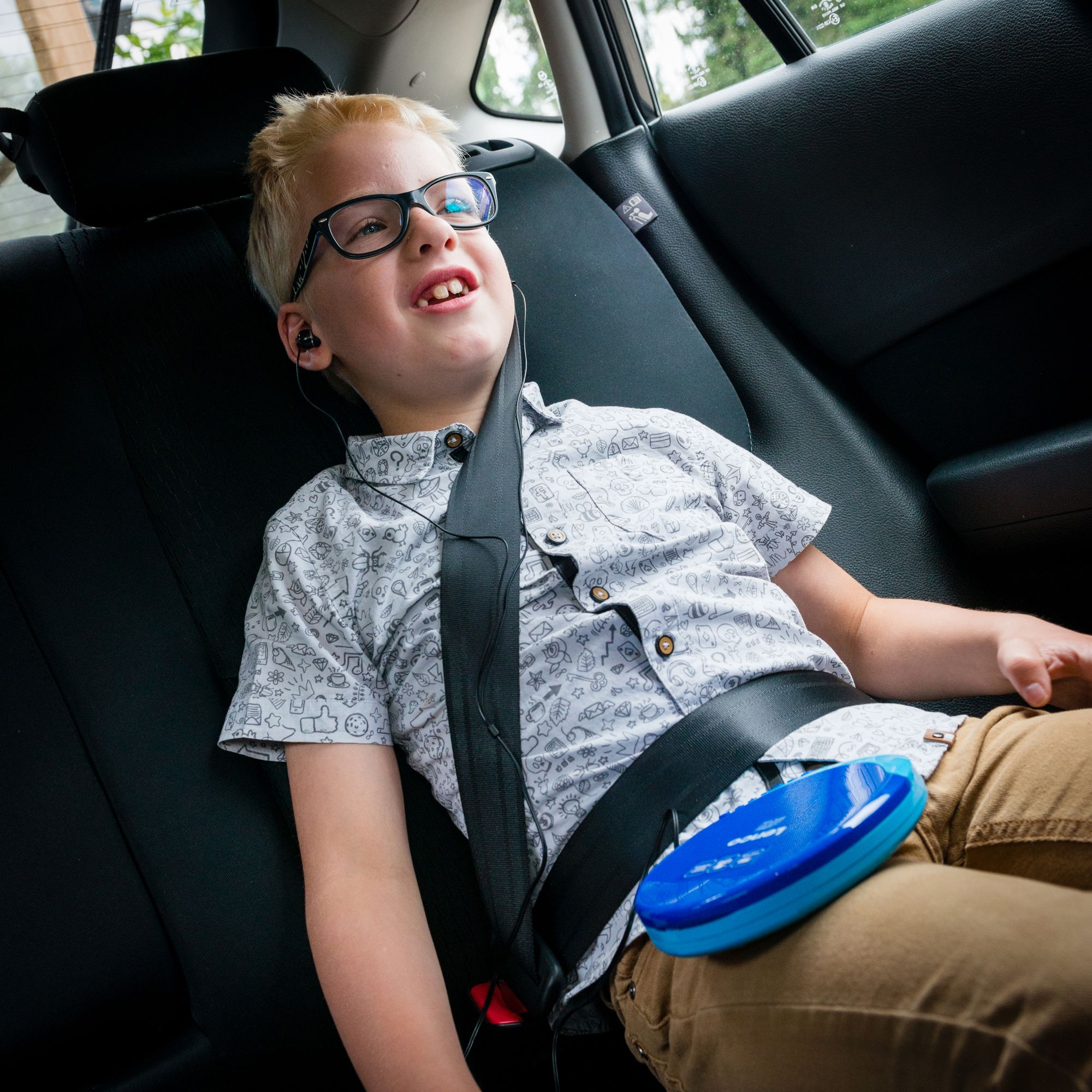 Chłopiec w foteliku samochodowym nosi okulary i pas bezpieczeństwa, ze słuchawkami. Niebieskie urządzenie leży na jego kolanach.