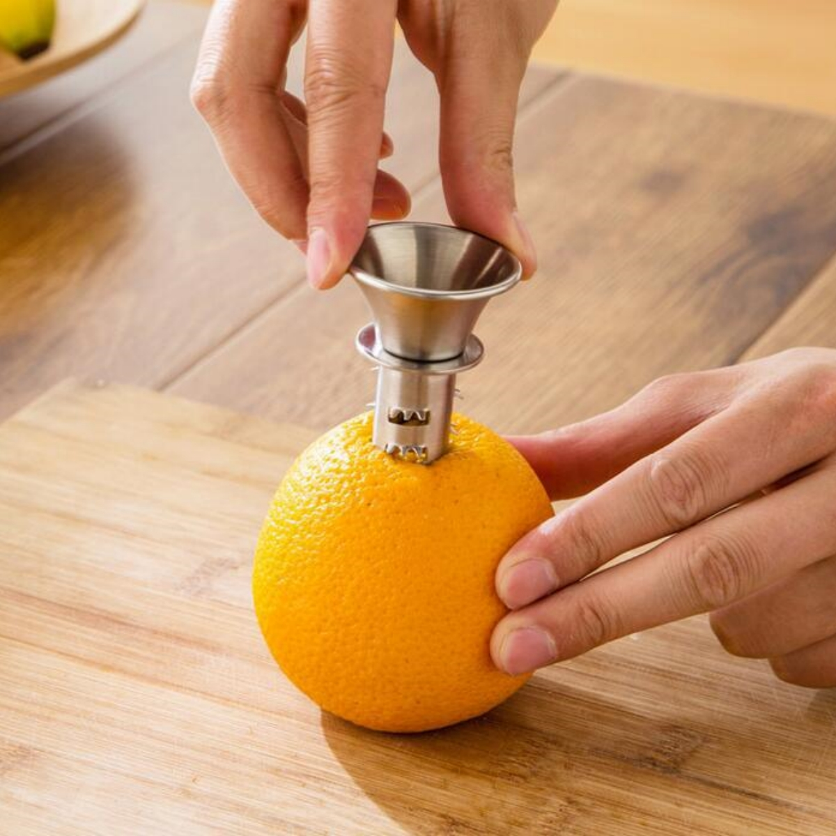 Hände, die einen Zitruspresser aus Edelstahl auf einer Orange verwenden. Holzschneidebrett im Hintergrund.