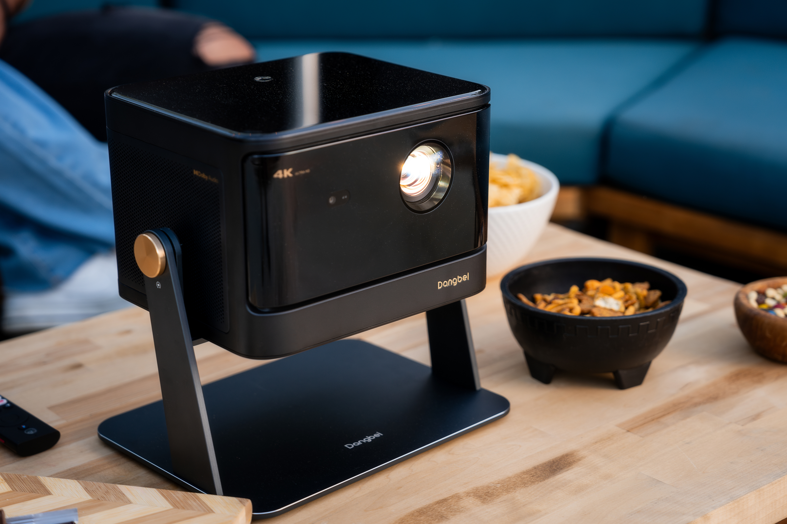 Czarny projektor na stole. Widoczne przekąski i pilot. Tłem jest otoczenie zewnętrzne.