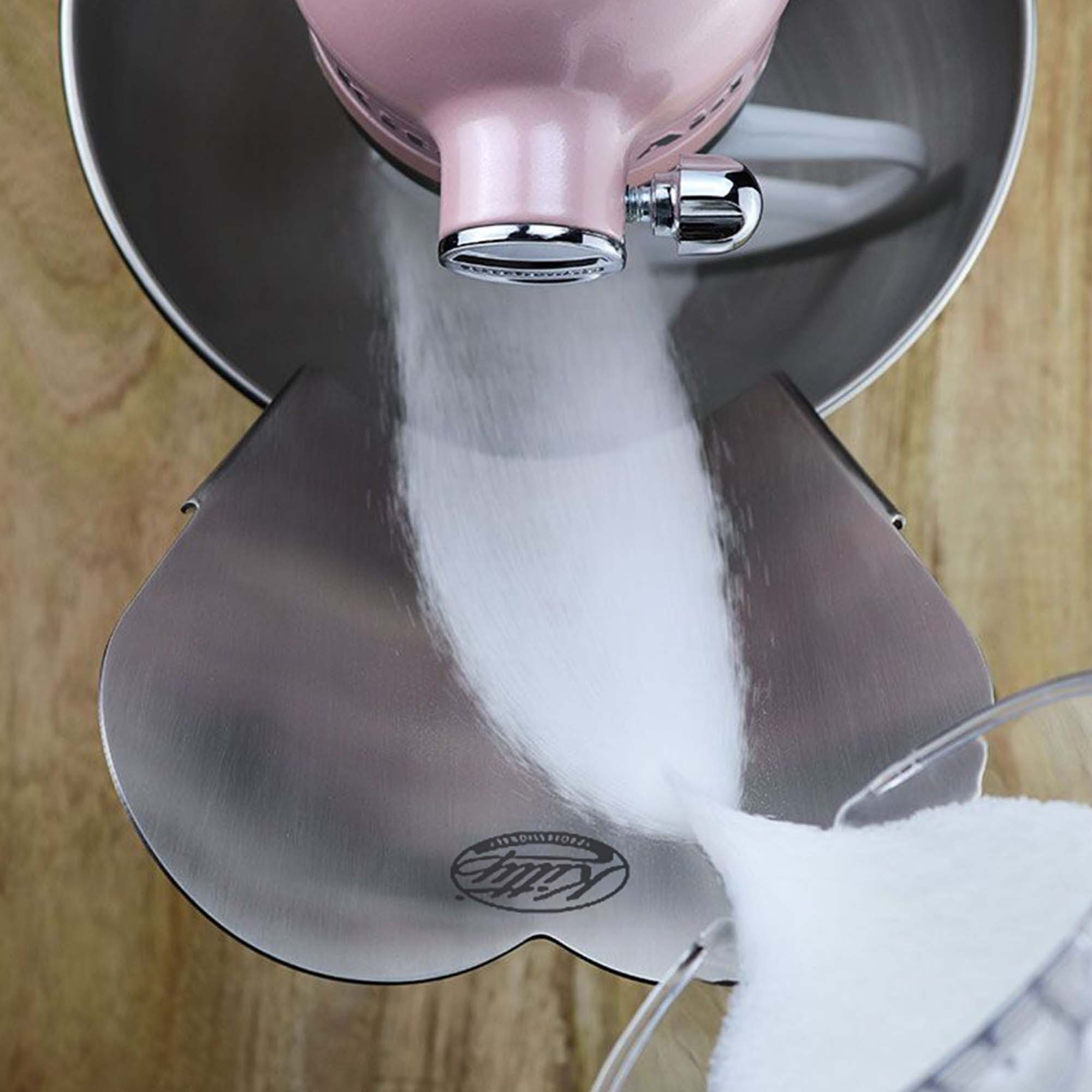 Ein rosa Küchenmixer mit weißem Zucker, der in eine Schüssel gegossen wird. Der Mixer steht auf Holz.