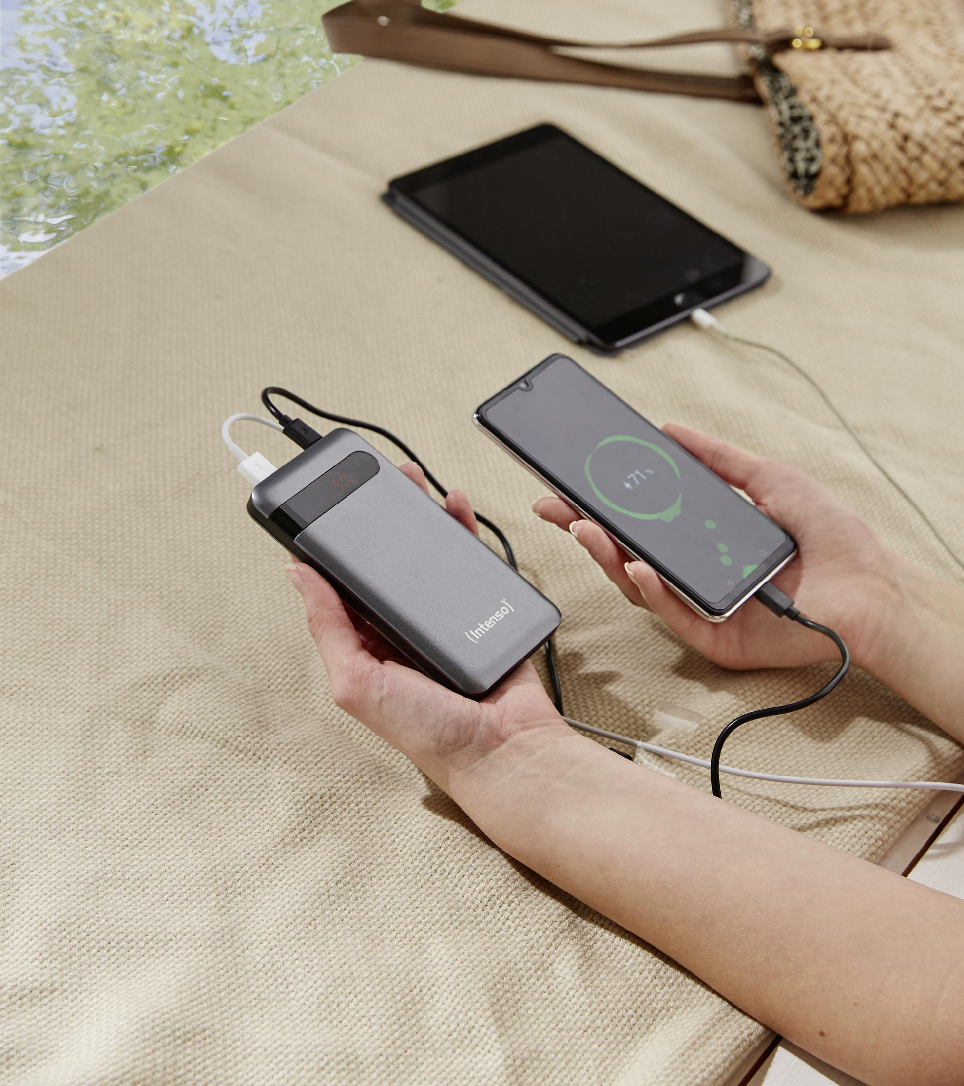 Hände halten eine graue Powerbank, die ein Telefon und ein Tablet auflädt. Im Hintergrund ist eine beige Oberfläche.