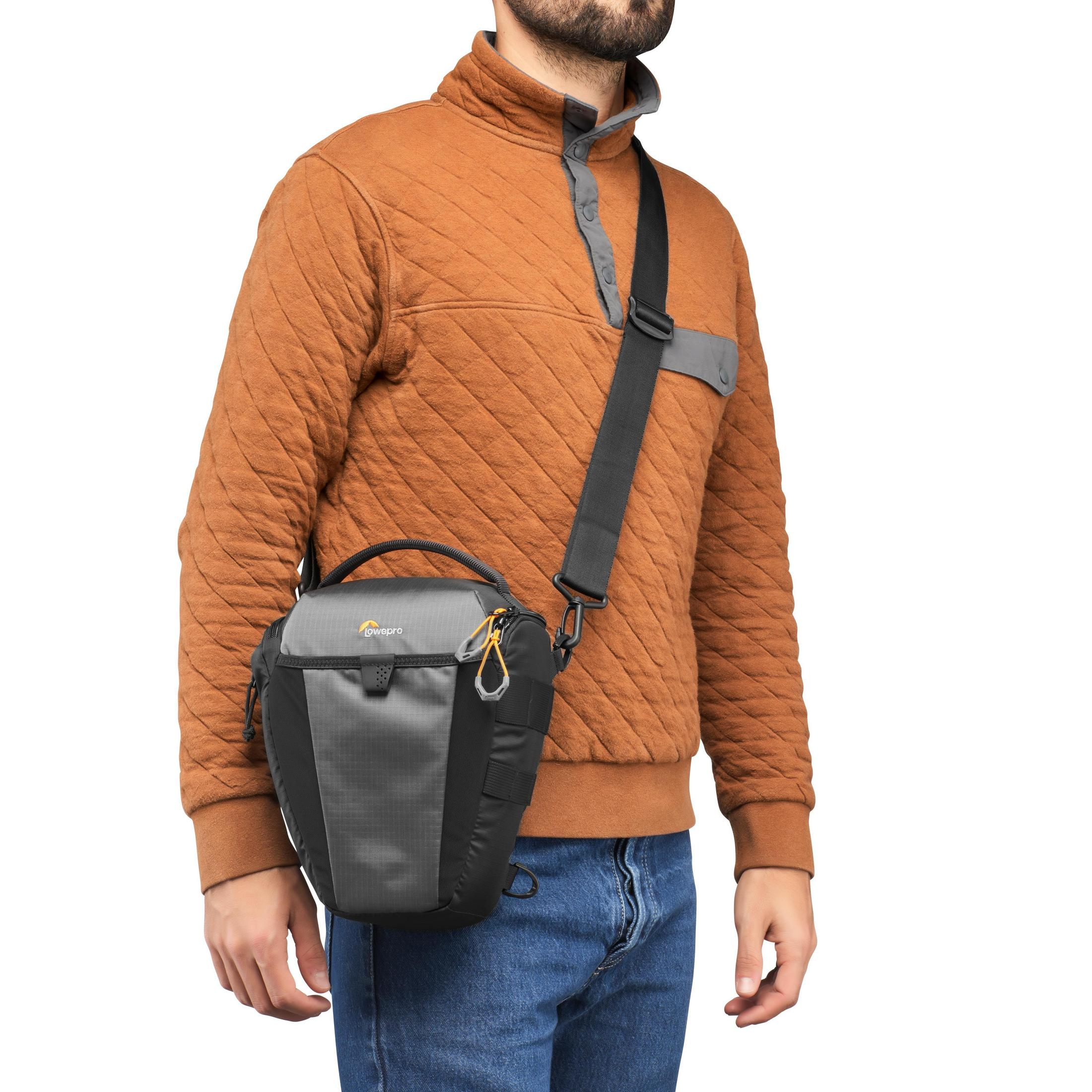 Un homme portant un pull marron et un sac photo gris sur l'épaule droite.