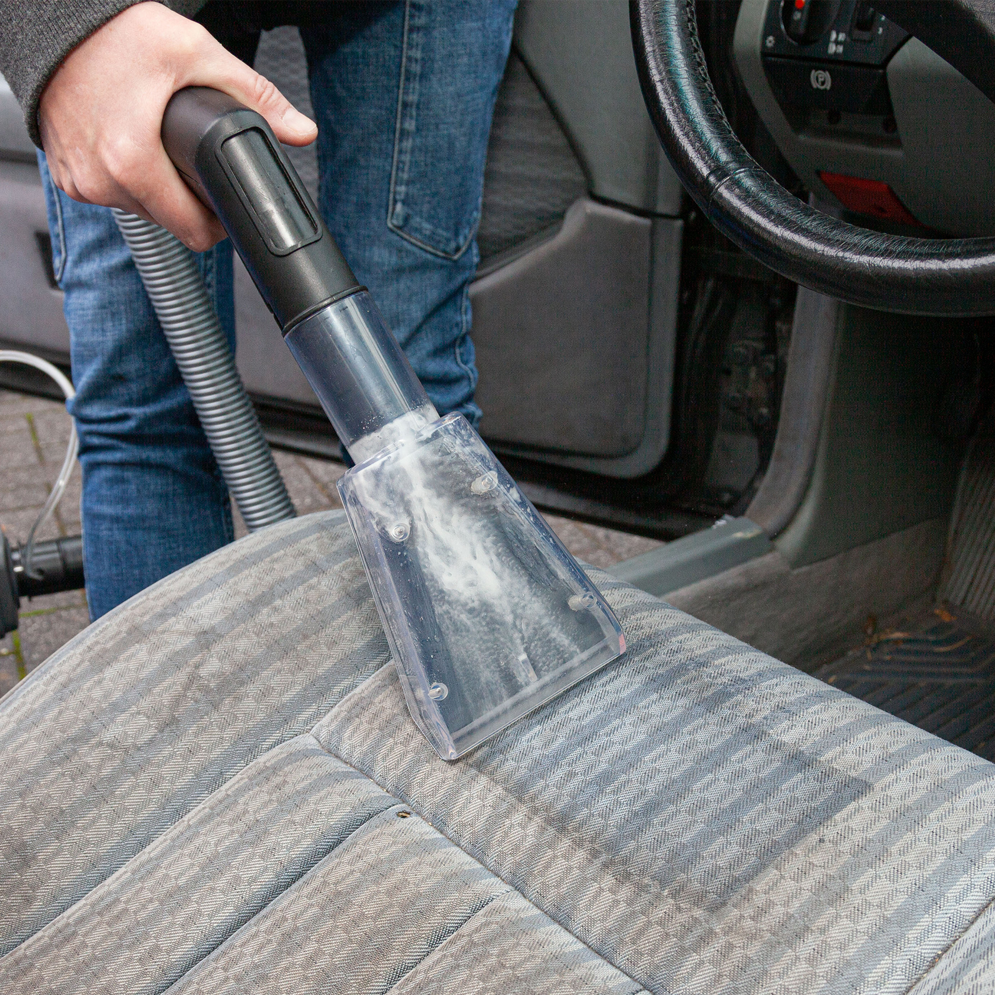 Person reinigt Autositz mit einem Staubsauger. Der Hintergrund ist ein Autointerieur.