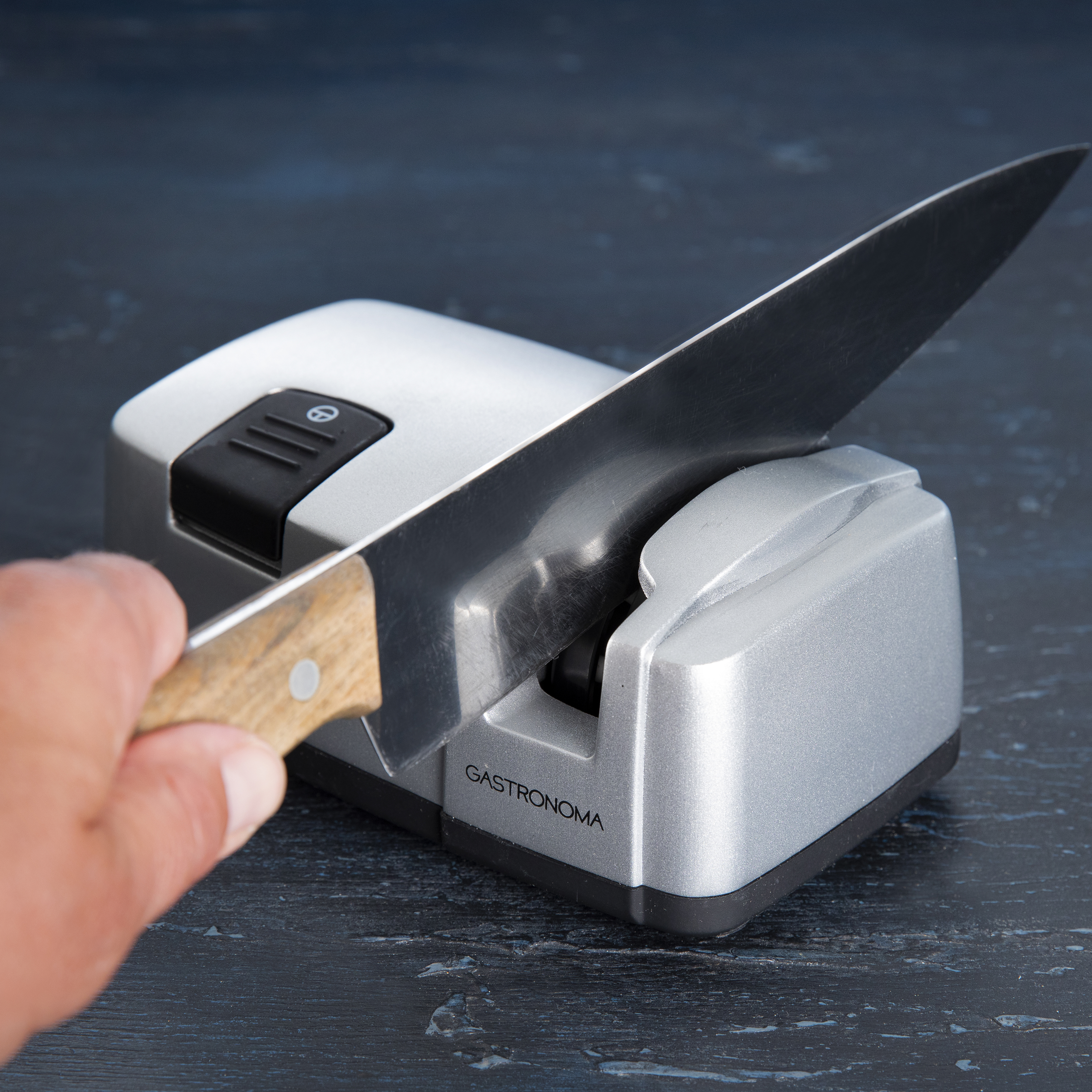 Ręka ostrzy nóż za pomocą szarej ostrzałki do noży Gastronoma. Ciemnoniebieskie tło.