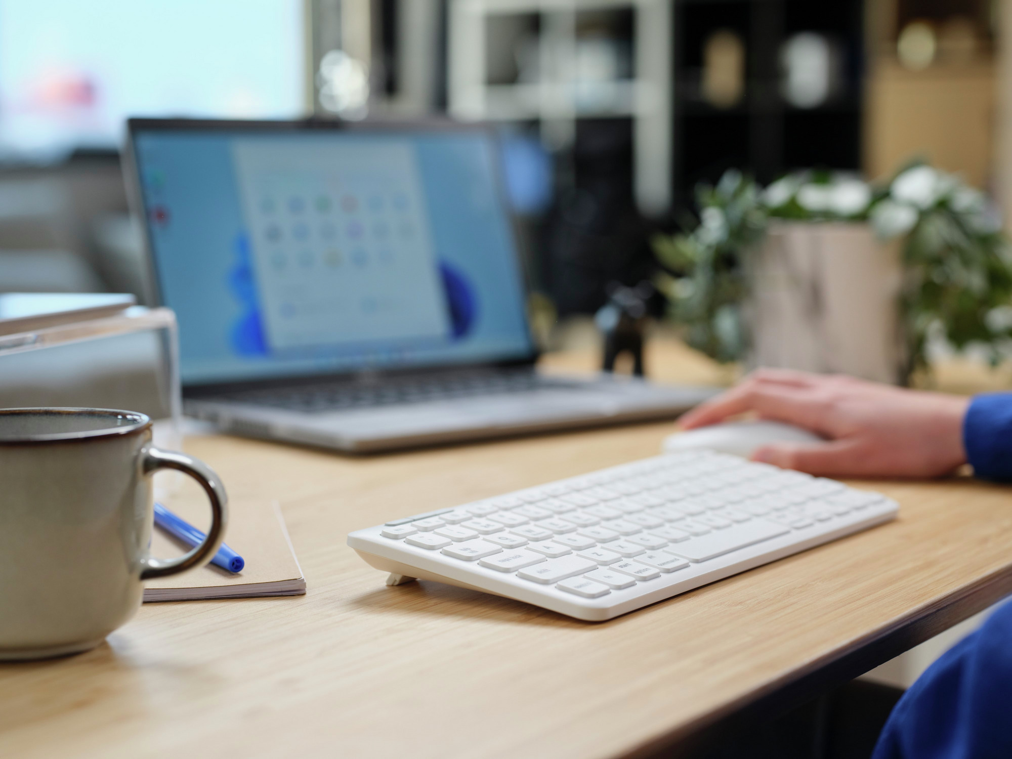 Bureau met laptop, toetsenbord, muis en een kopje. Een hand gebruikt de muis.