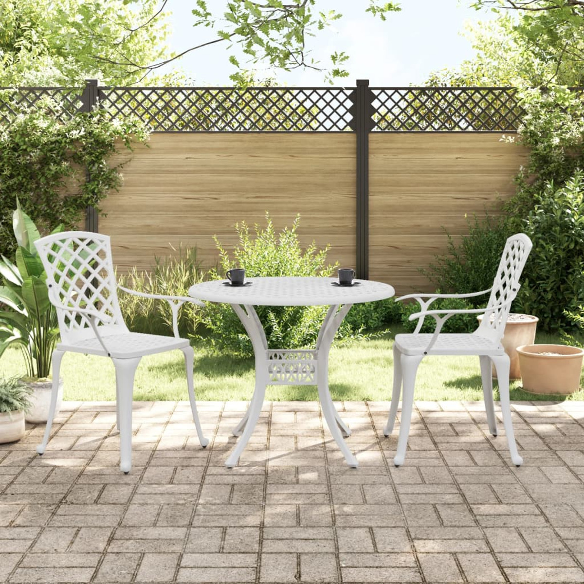Ein weißes Terrassenset mit Tisch und zwei Stühlen auf einer Ziegelterrasse, mit Grün und Zaun.