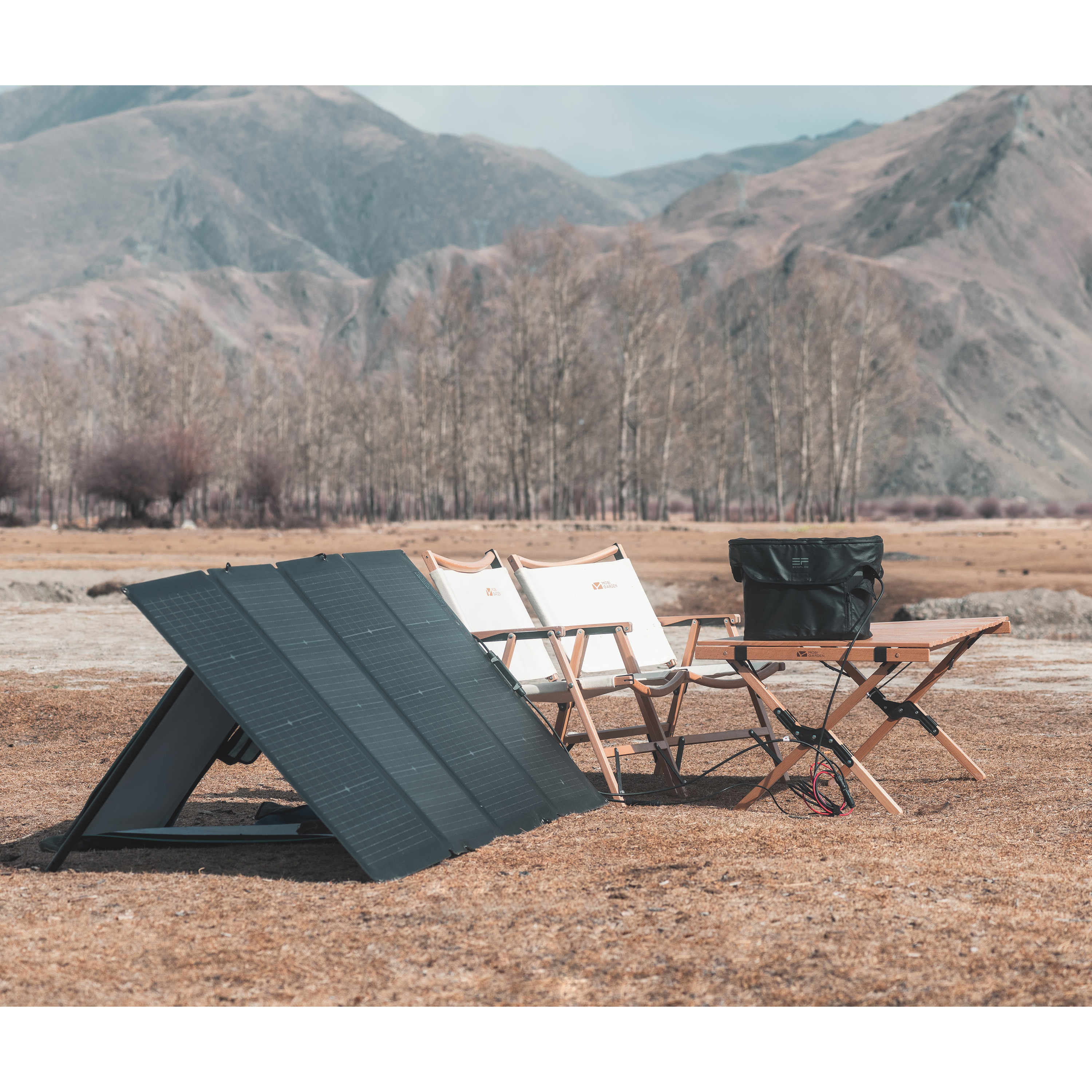 Scena na zewnątrz. Panele słoneczne, składane krzesła, stół i torba przed górami.