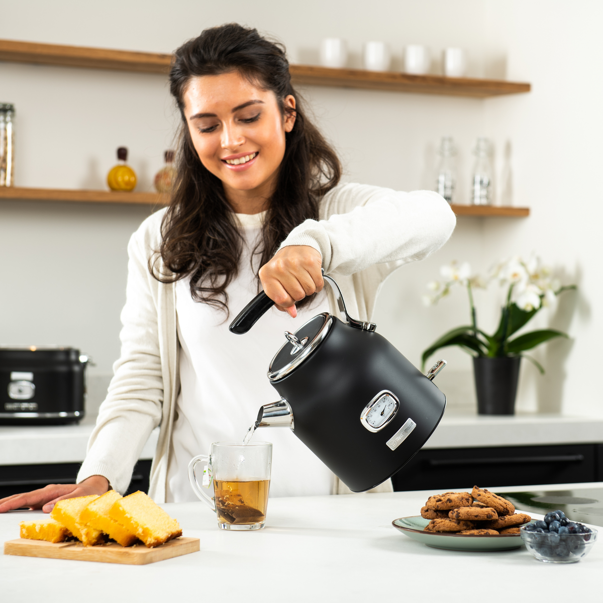 Frau gießt heißes Wasser aus einem schwarzen Wasserkocher in eine Teetasse, mit Essen auf dem Tisch.