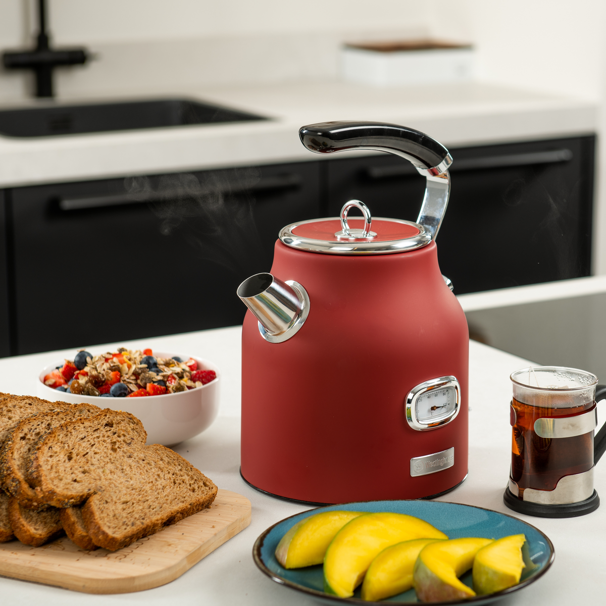 Roter Wasserkocher auf weißem Tisch mit Frühstück: Brot, Obst und eine Tasse Tee.