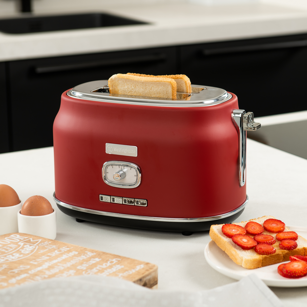 Roter Retro-Toaster mit zwei Toastscheiben auf einer weißen Arbeitsplatte, Eier im Hintergrund.