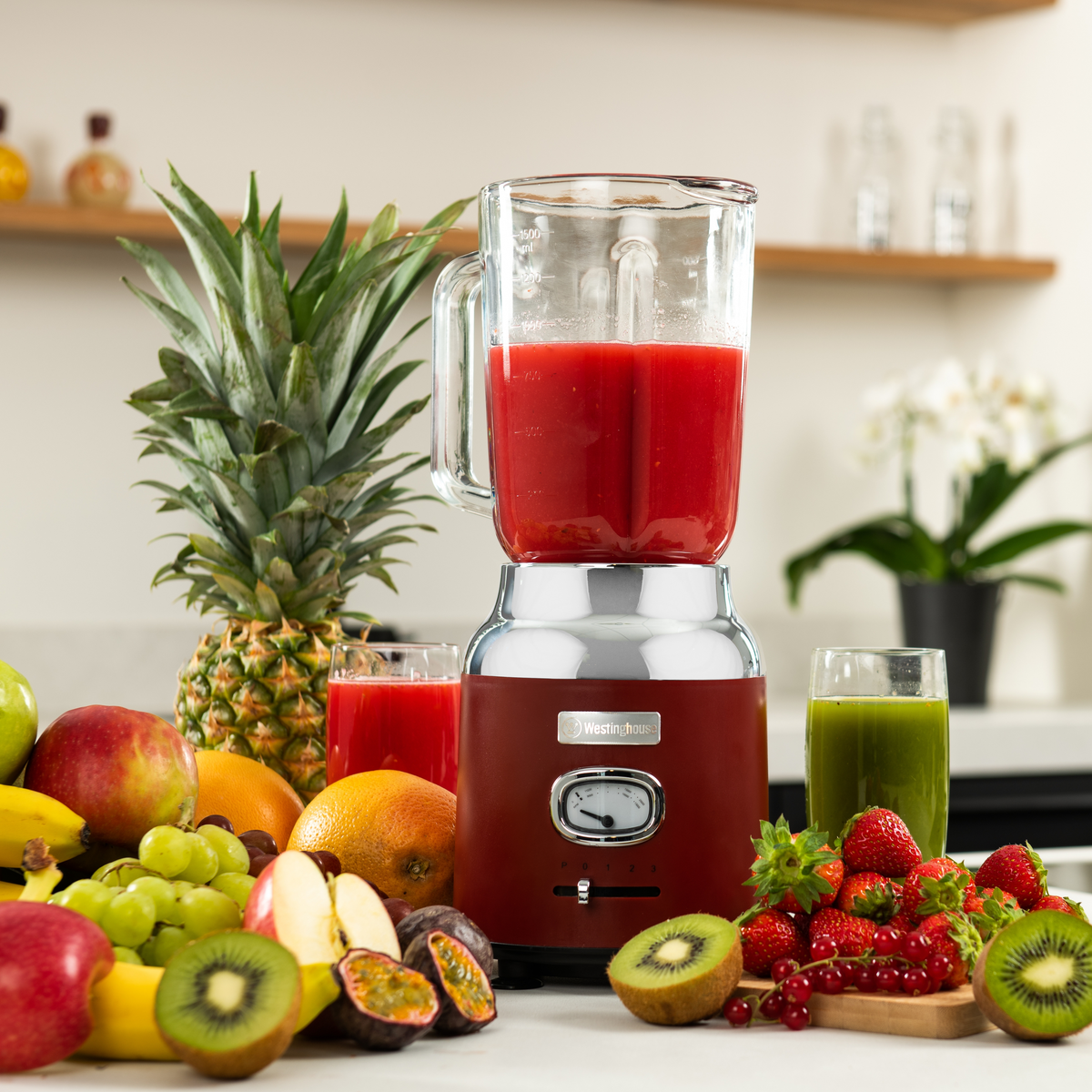 Mixer mit roter Flüssigkeit, Früchten und Saftgläsern auf einem Tisch. Ananas und Kiwi sind sichtbar.