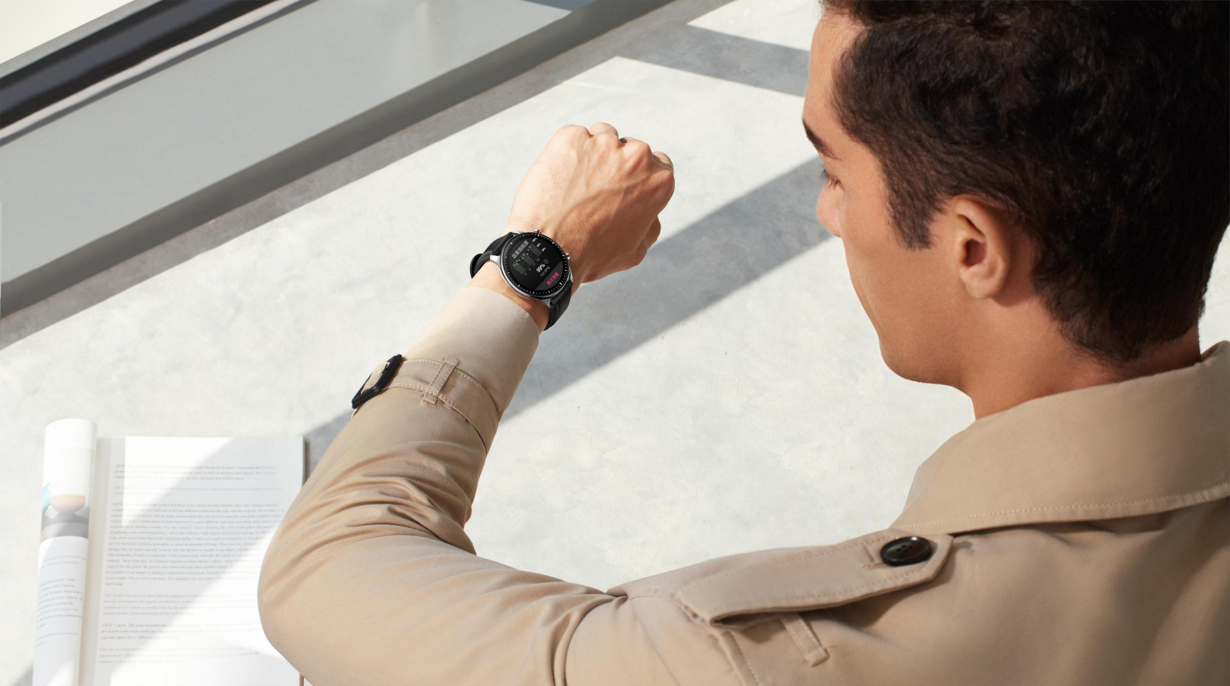 Ein Mann in beigem Mantel betrachtet seine schwarze Smartwatch. Er ist an einem Fenster mit Schreibtisch.