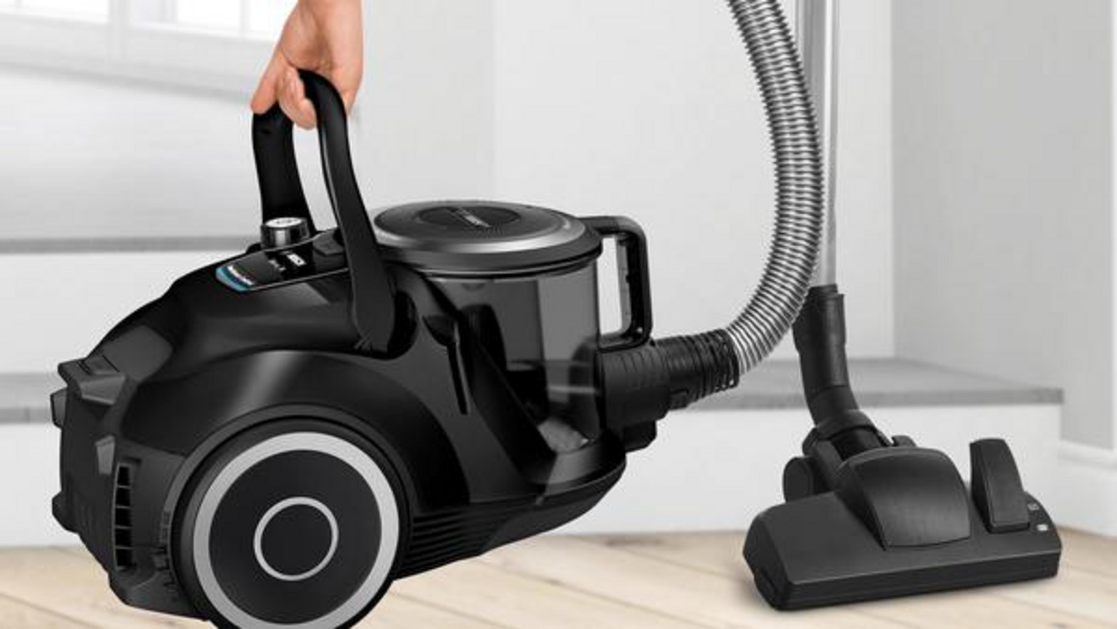 Czarny odkurzacz trzymany przez rękę. Ma wąż i stoi na drewnianej podłodze.