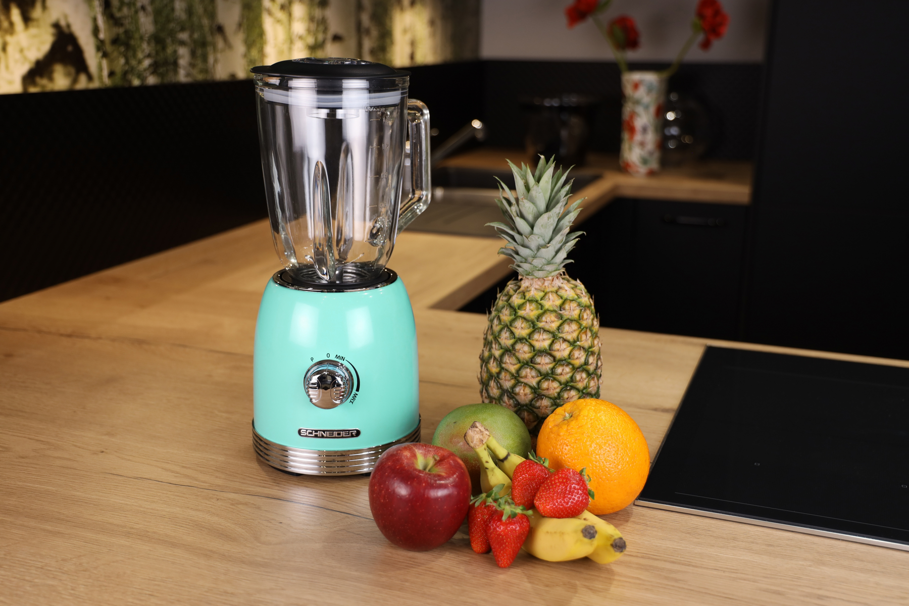 Ein Mixer mit Früchten auf einer Holztheke. Enthält Ananas, Apfel, Orange, Mango und Erdbeeren.