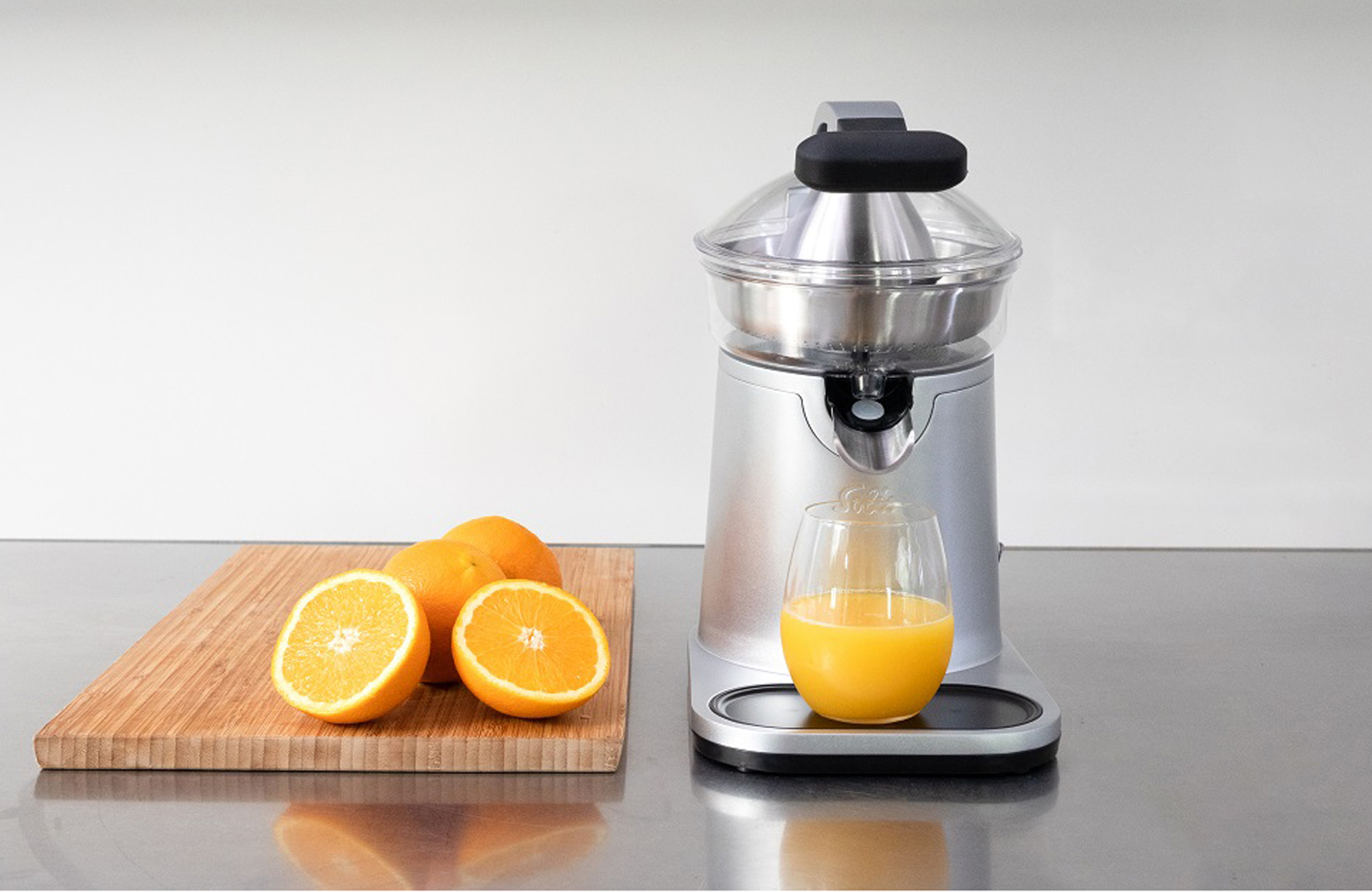 Silberne Saftpresse mit Orangen auf einem Holzbrett und ein Glas Saft auf Edelstahl-Arbeitsplatte.