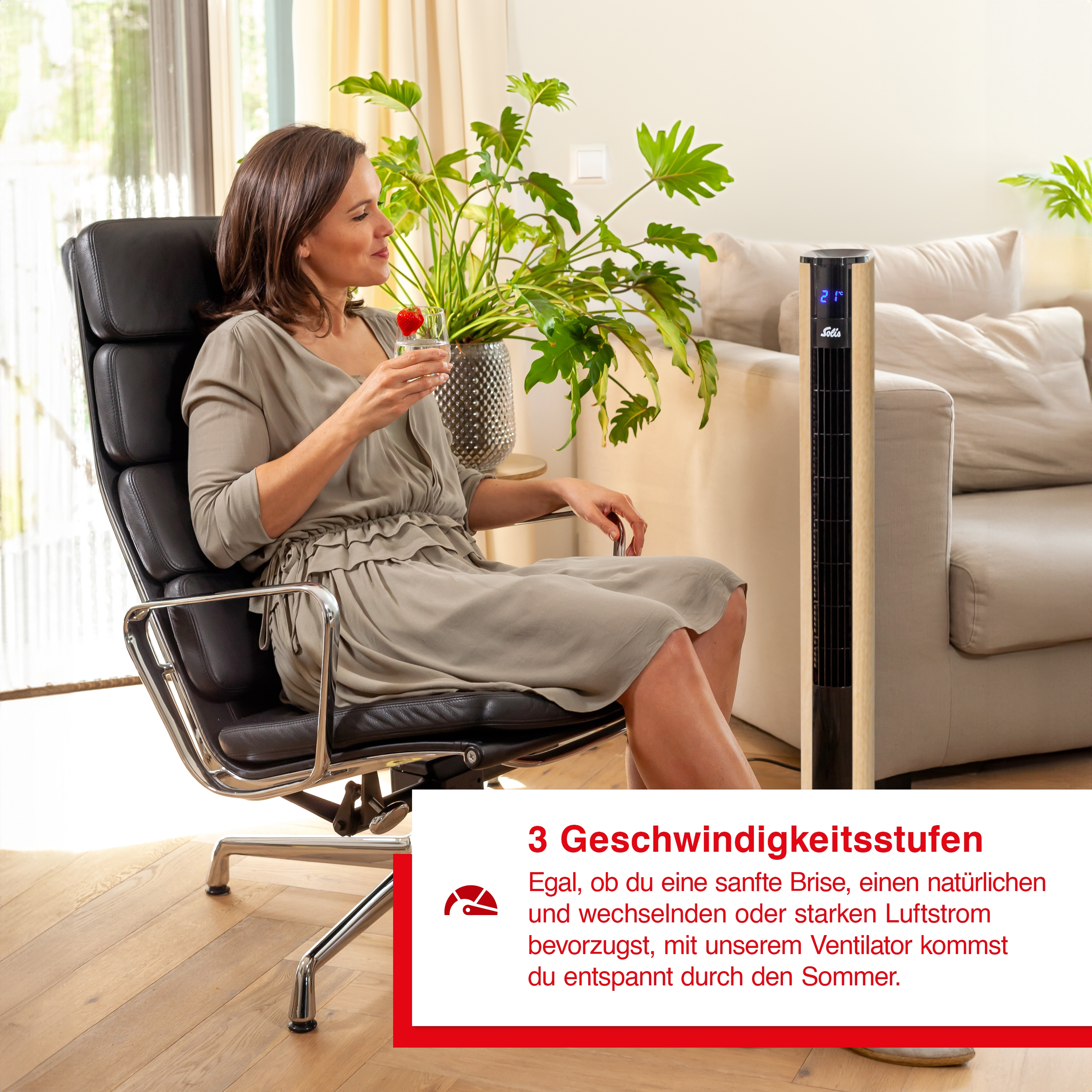 Frau im Stuhl, trinkend; ein hoher Ventilator und eine Pflanze sind in der Nähe.
