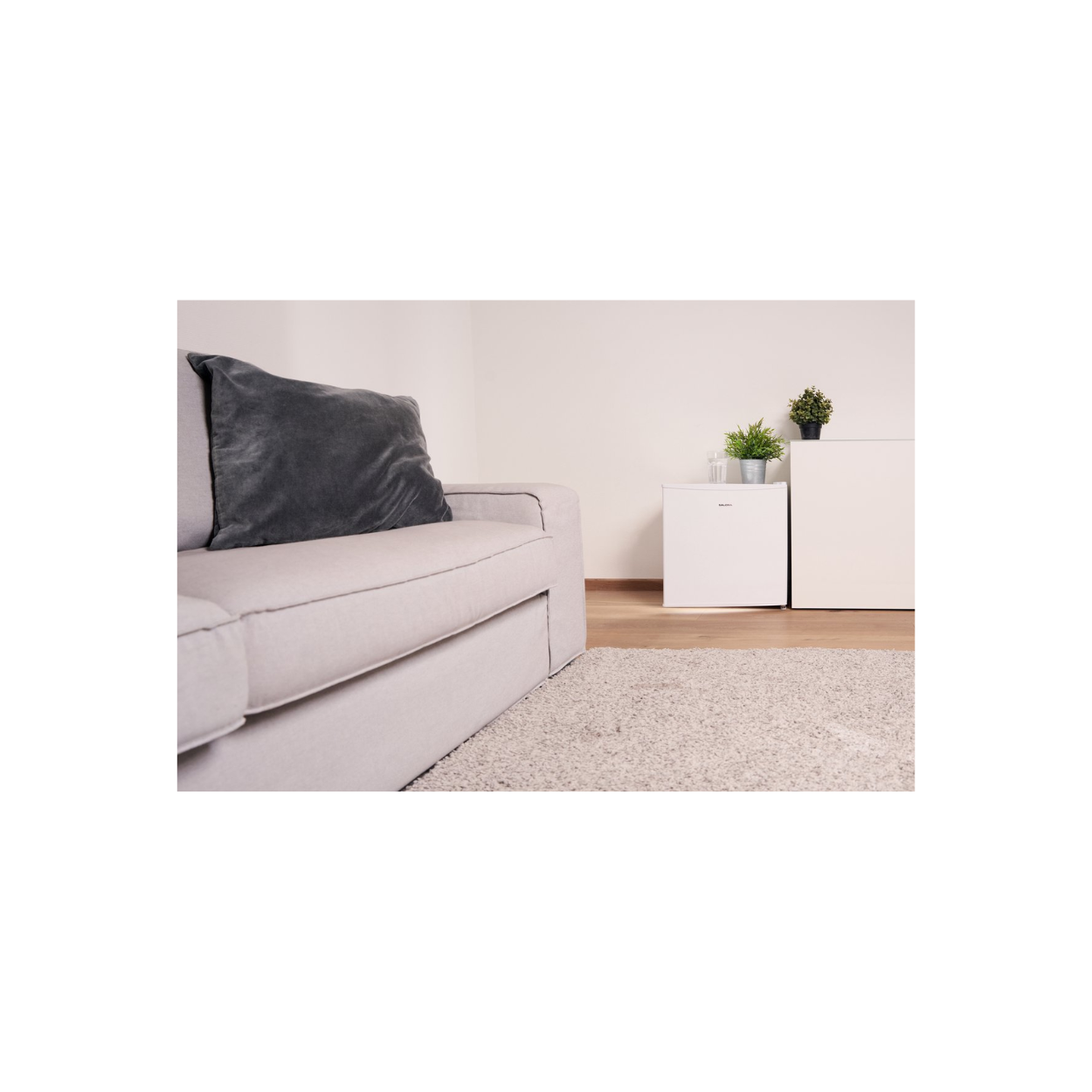 Scène de salon avec canapé gris, tapis et mur blanc. Un mini-réfrigérateur blanc est visible.