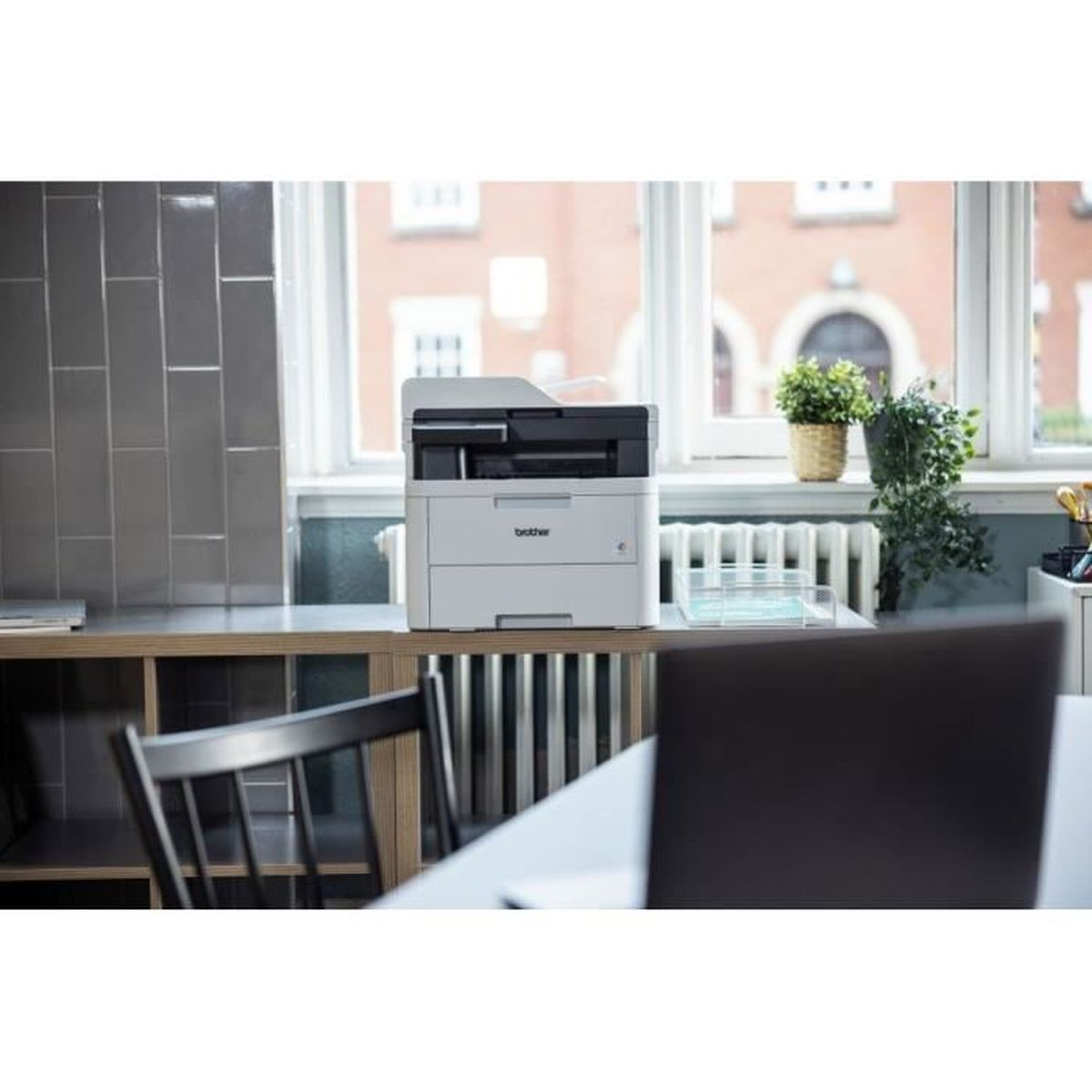 Drukarka Brother stoi na półce, w pobliżu okna z widokiem na ceglany budynek.