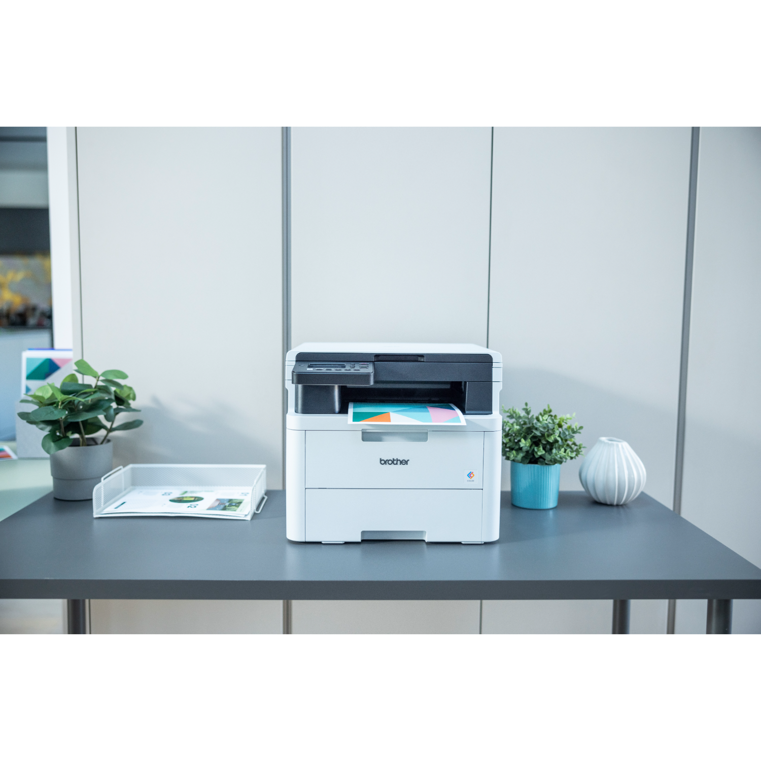 Czarno-biała drukarka na szarym biurku. W pobliżu znajdują się rośliny i inne artykuły biurowe.