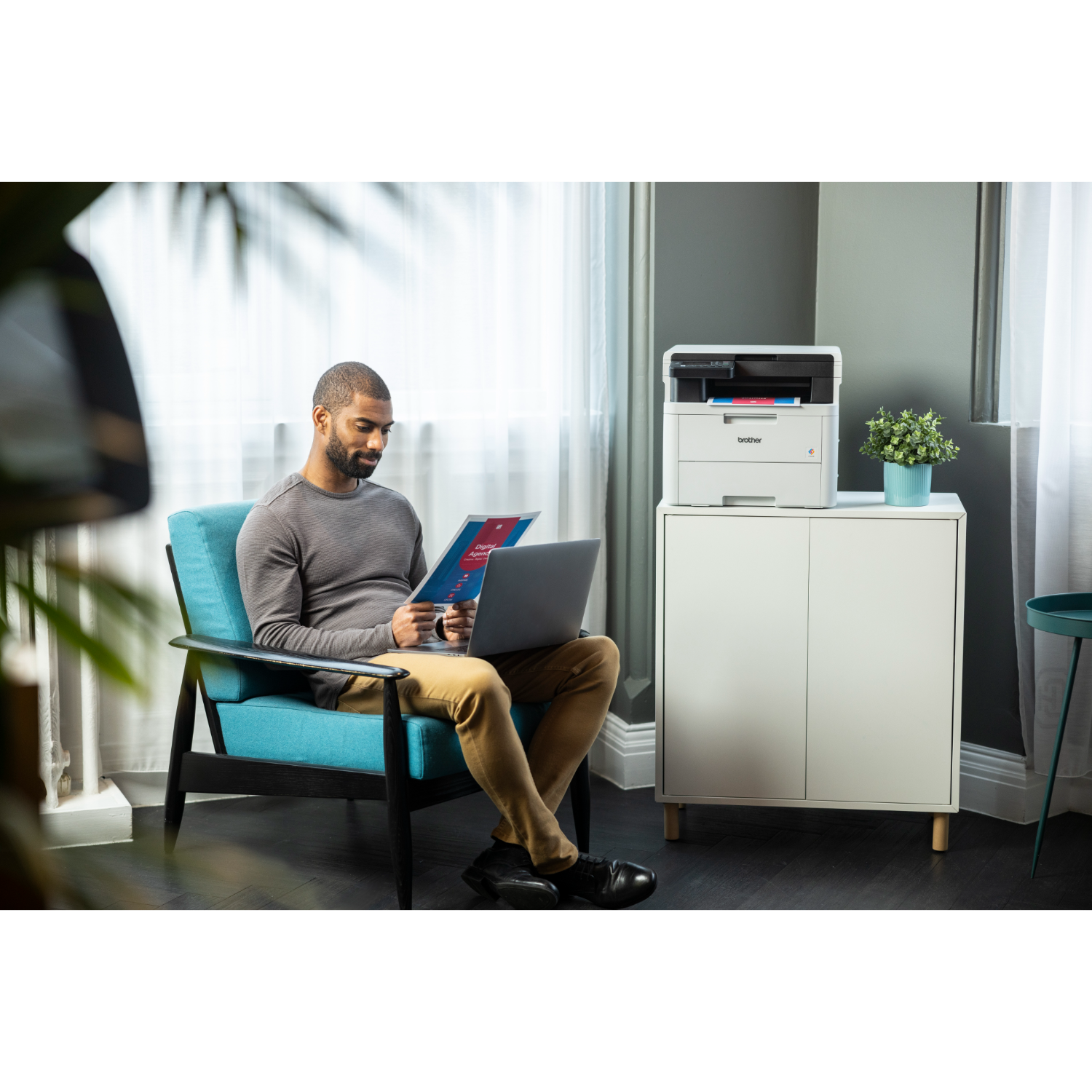 Mężczyzna siedzi na niebieskim krześle, używając laptopa i przeglądając dokument. Drukarka i rośliny są w pobliżu.