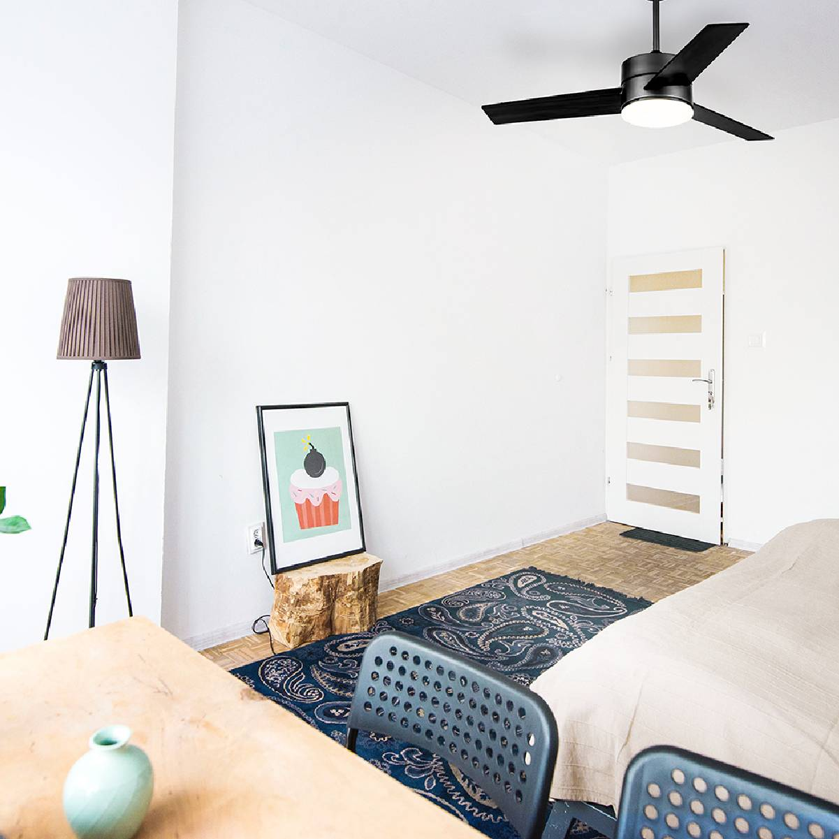 Una stanza con pareti bianche, un ventilatore a soffitto e un'immagine di un cupcake. Lampada e letto visibili.