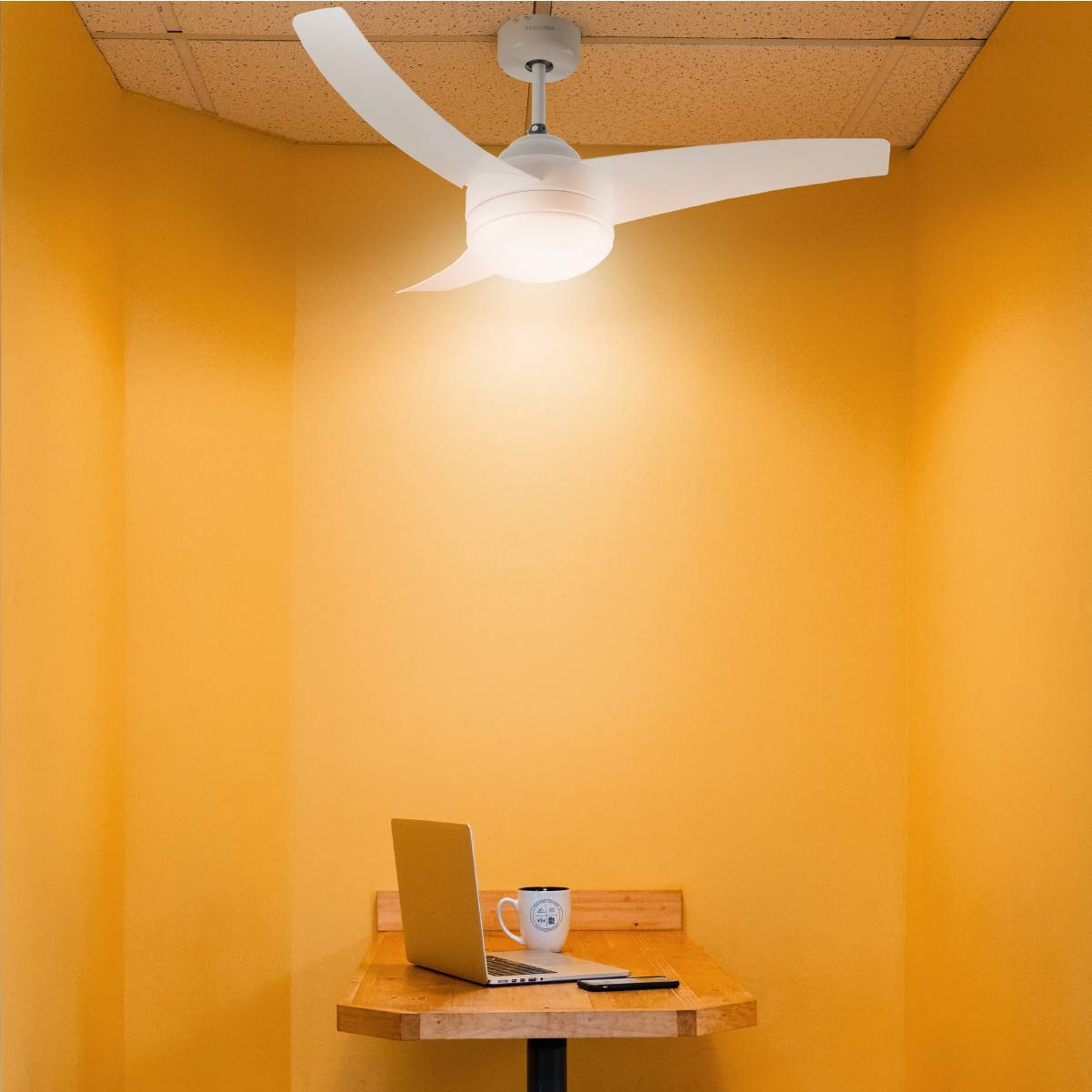 Stanza con pareti gialle con ventilatore a soffitto bianco, laptop e tazza su un tavolo di legno.